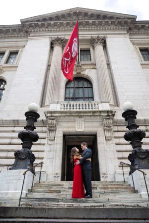 Couple embracing on steps of neoclassical building under a red flag in New York City.