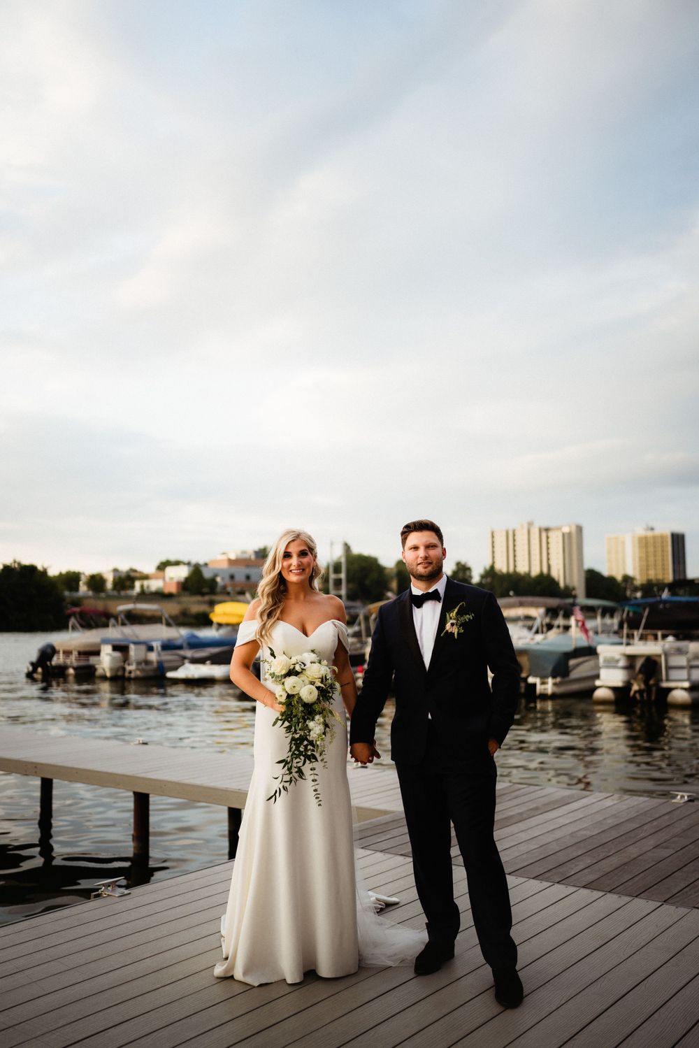 A sunset wedding portrait on wooden dock with boats and city skyline in distance.