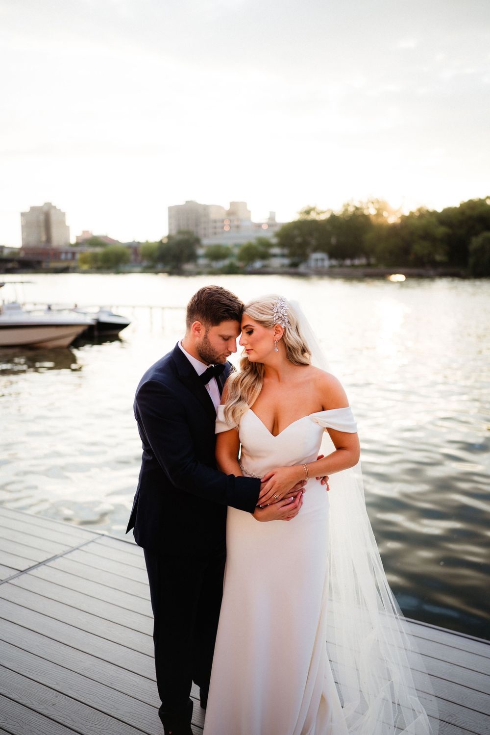 A romantic sunset waterfront wedding portrait on a wooden dock with boats and cityscape in background.