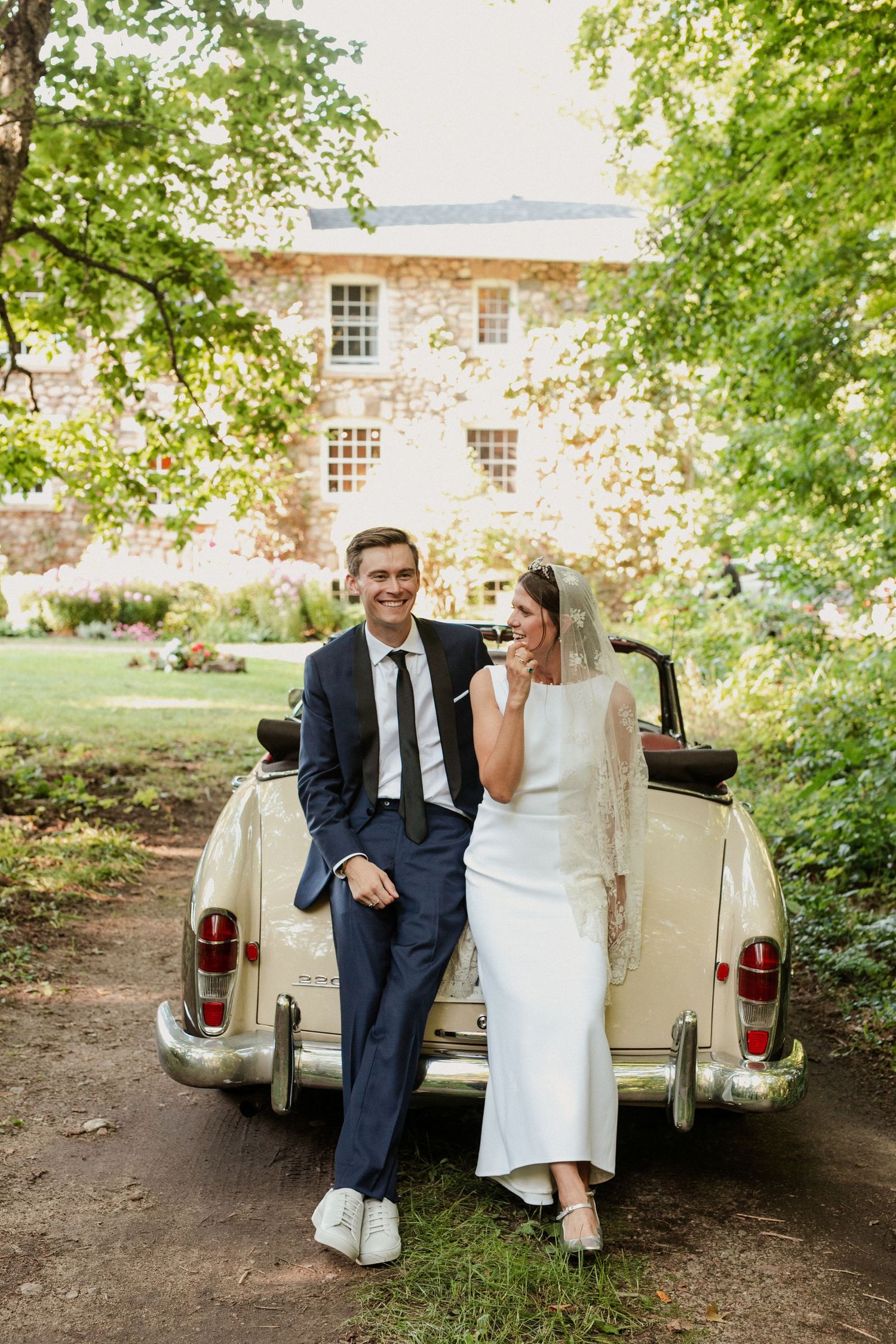 Bride and groom leaning against vintage cream convertible car in lush garden setting.