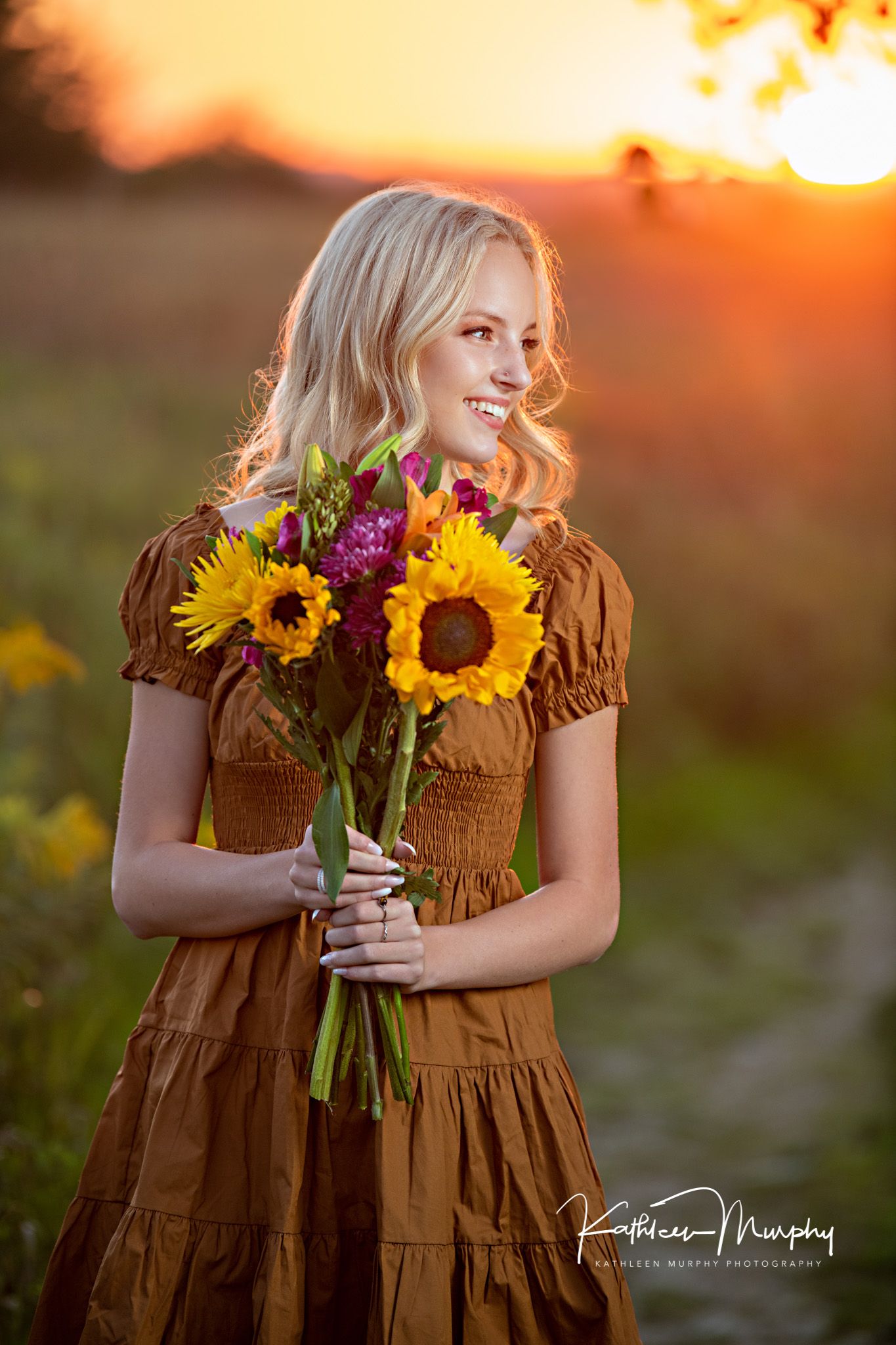 Golden sunset portrait holding vibrant sunflower and purple flower bouquet in brown dress.