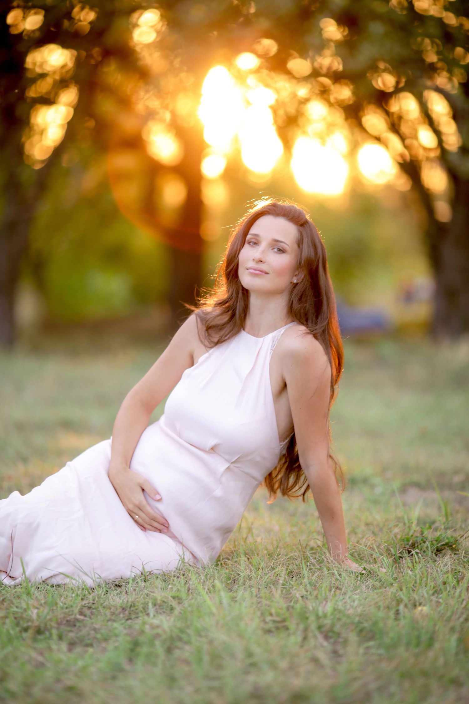 Maternity photo of woman in flowing white dress reclining on grass during sunset.