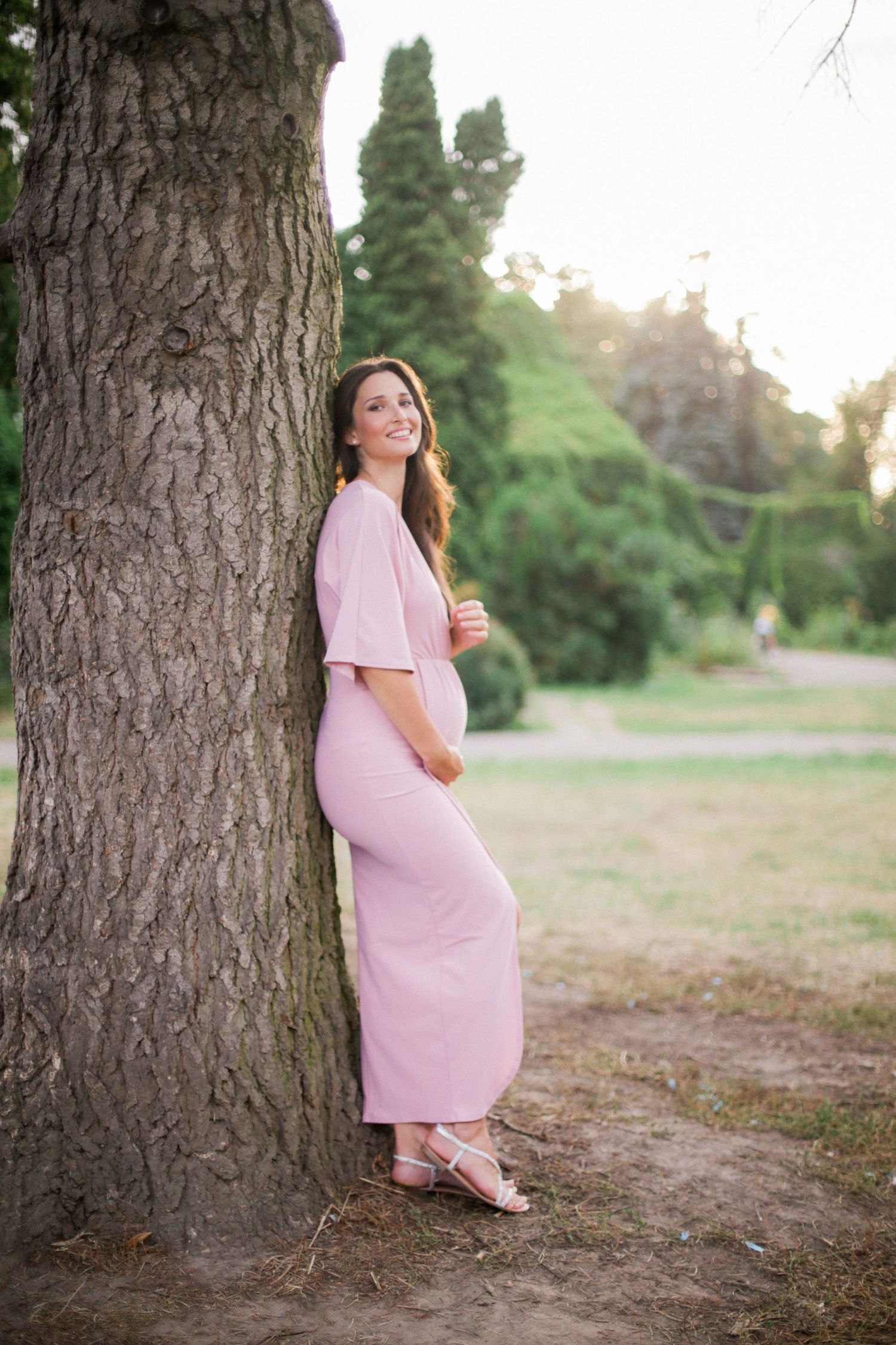 Maternity portrait in pink dress leaning against large tree trunk.