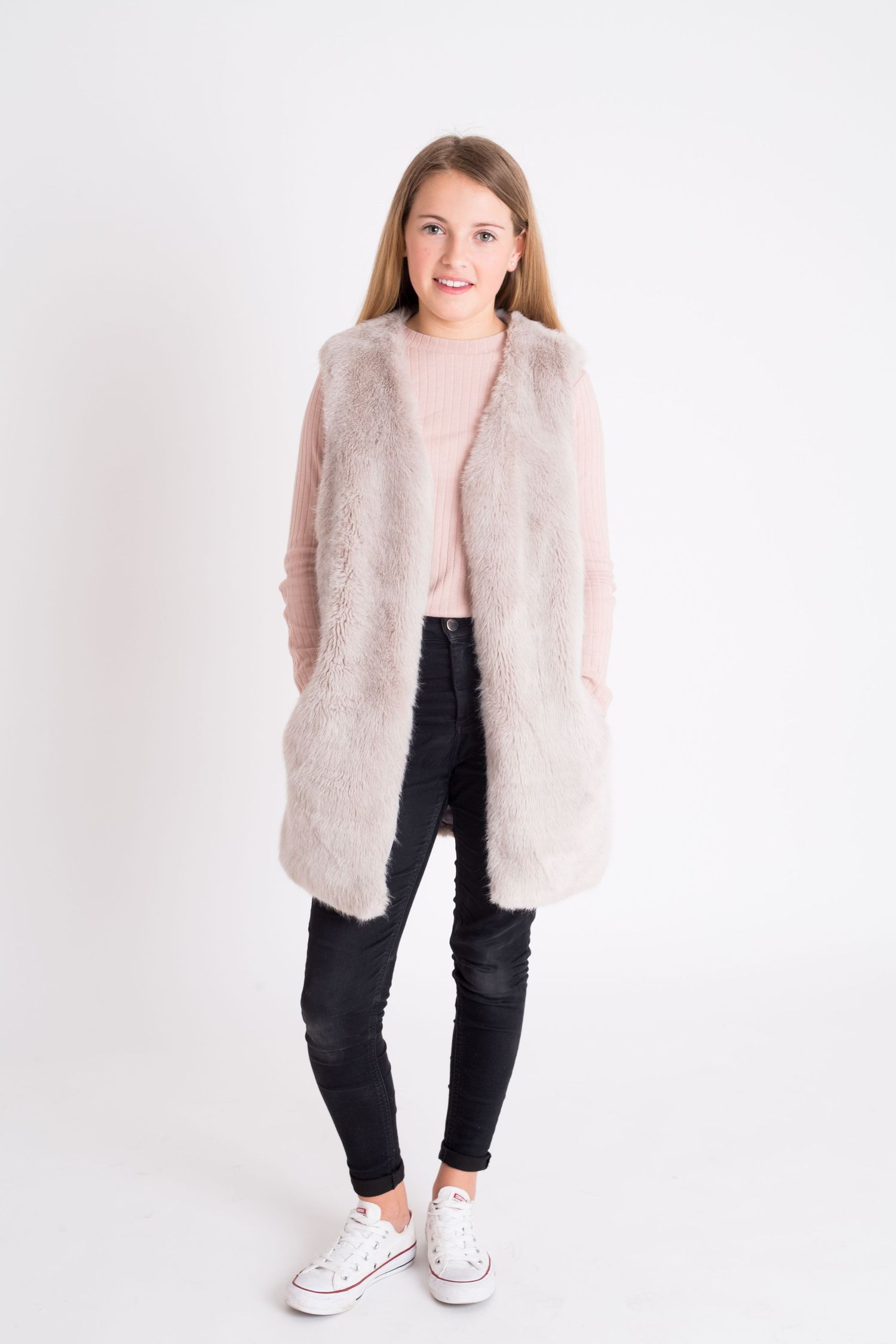 Person wearing pink fuzzy vest with black jeans and white sneakers against white background.