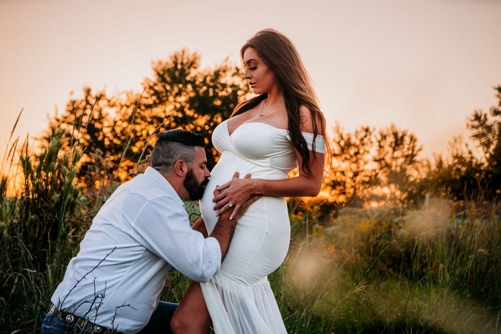 A maternity photo in a field at sunset shows an intimate moment while wearing a white dress.