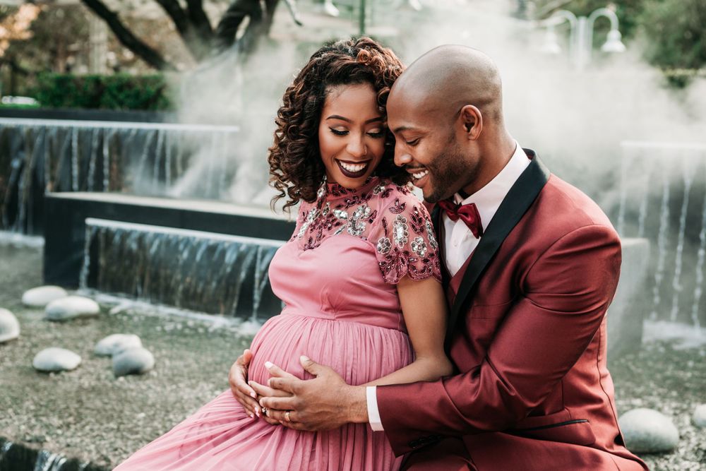 A couple shares an intimate moment by a waterfall, with the person in a pink dress cradling their pregnant belly.