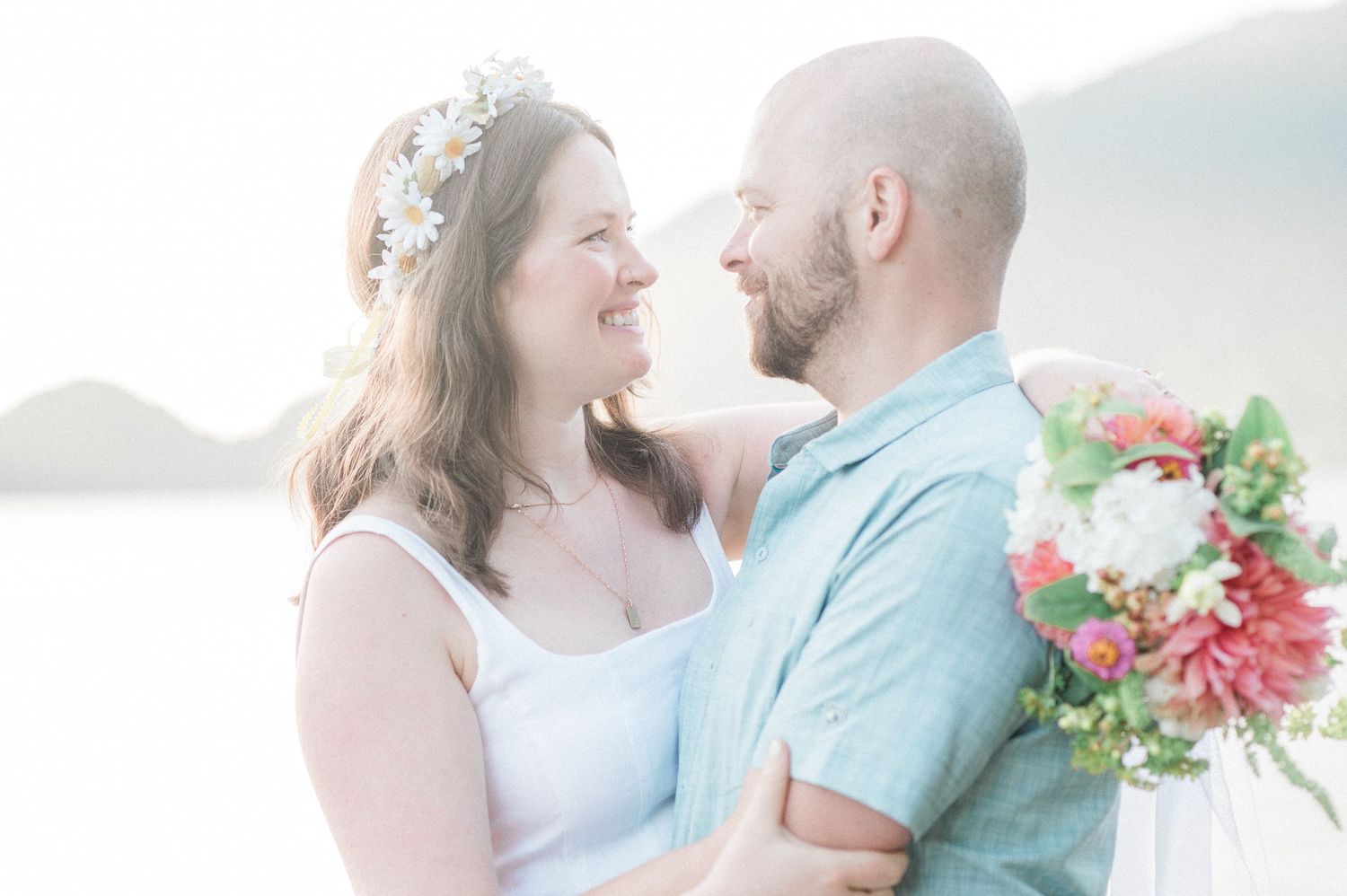 A romantic couple embraces on a misty mountain overlook while holding a pink floral bouquet.