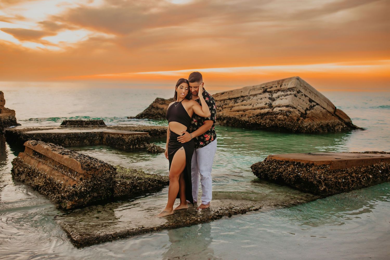 Couple sharing romantic moment on rocky beach formation during vibrant orange sunset.