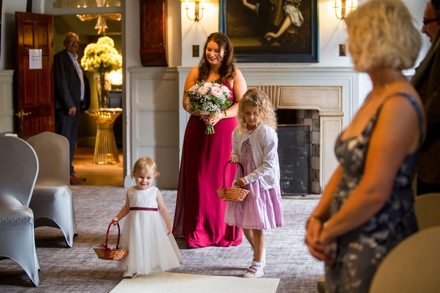 Bridesmaid in red gown walking down aisle with flower girls during wedding ceremony.