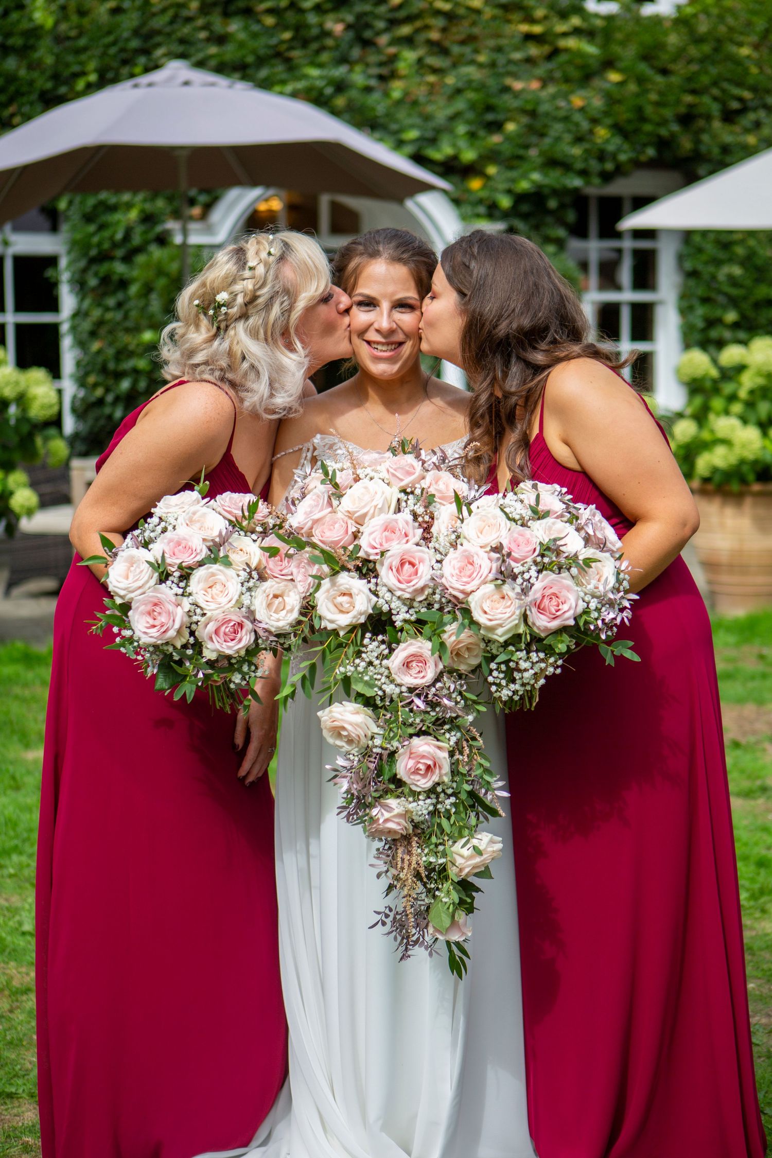 Bride in white gown holding pink roses bouquet, flanked by two bridesmaids in burgundy dresses kissing her cheeks.