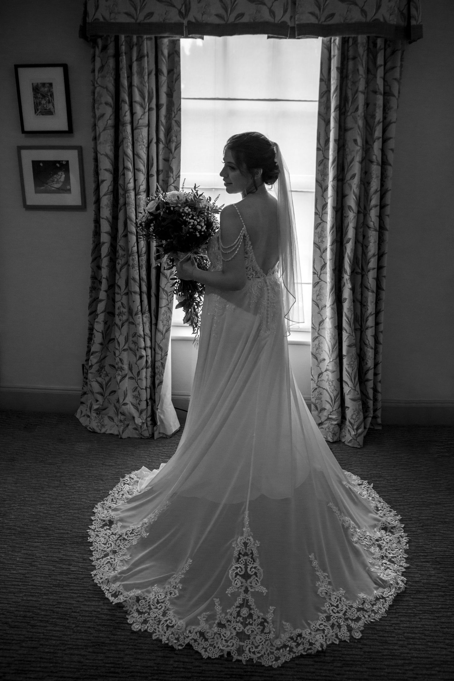 Elegant black and white portrait of bride in lace gown holding bouquet by window.