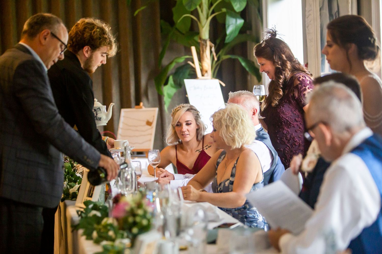 Wedding guests mingling and signing documents at a floral-decorated reception dinner table.