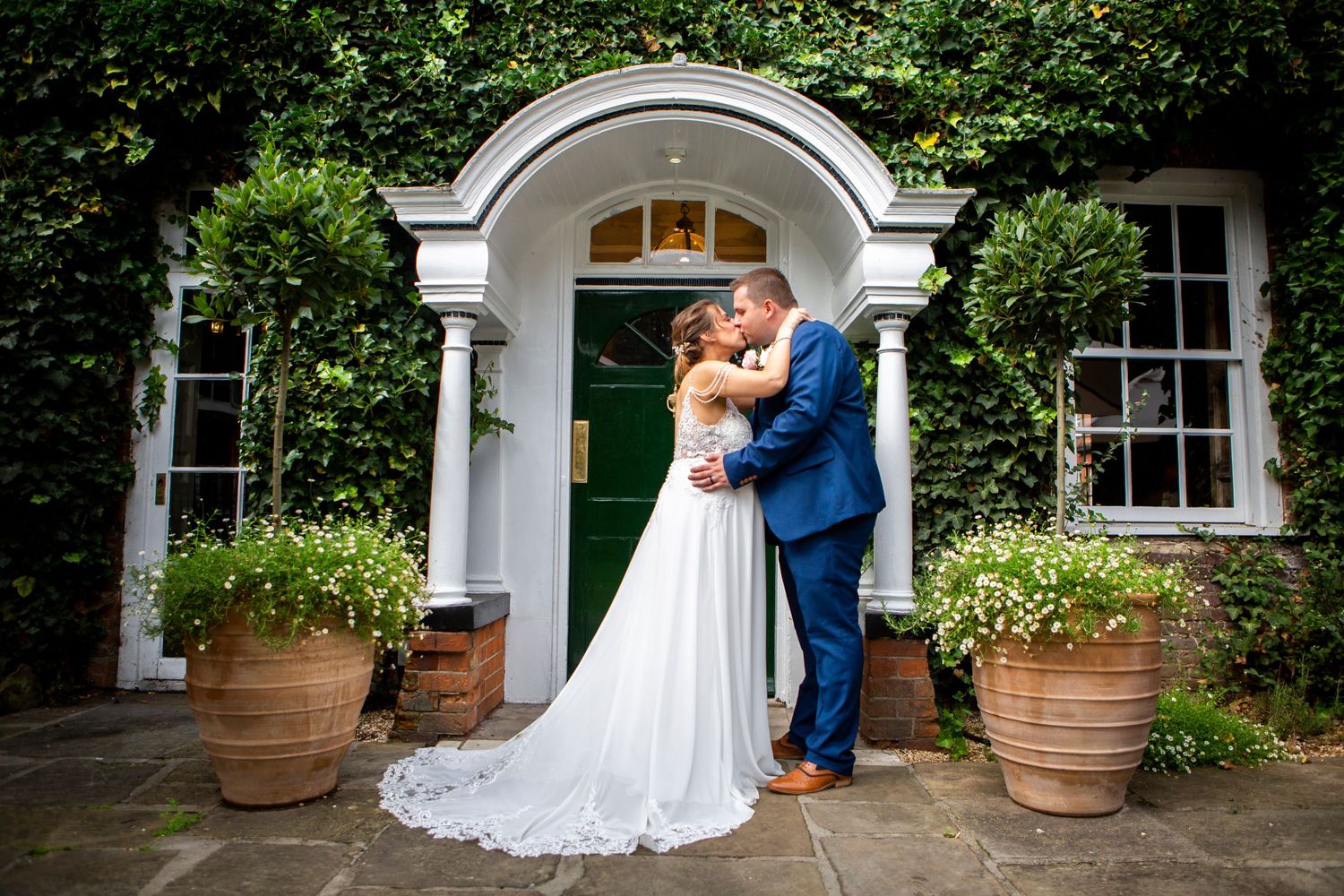 Bride and groom share romantic kiss outside ivy-covered wedding venue entrance with potted plants.