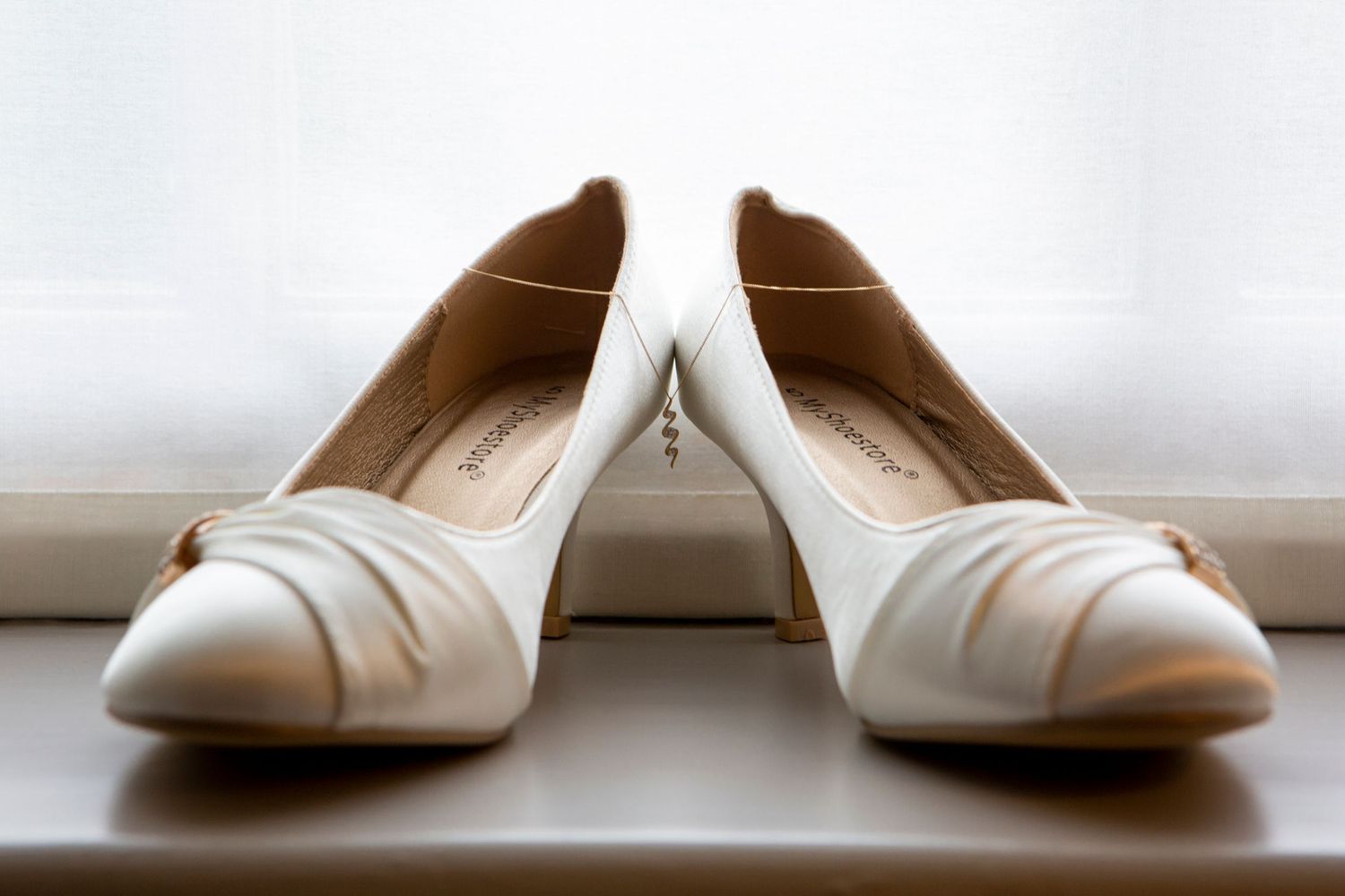 Pair of white satin bridal heels with bow detail displayed on a windowsill.