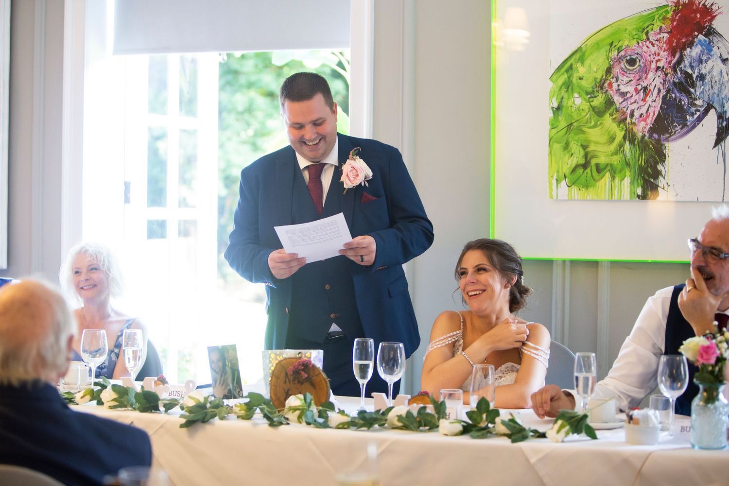 Groom delivering heartfelt wedding speech while bride smiles at elegantly decorated reception table.