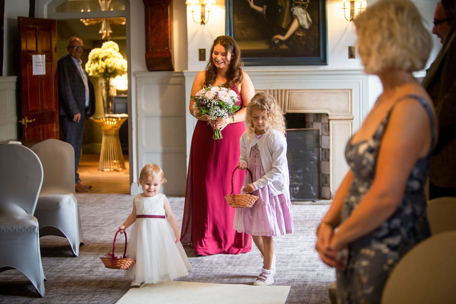 Bridesmaid in red gown walks with flower girls down aisle in elegant wedding ceremony venue.