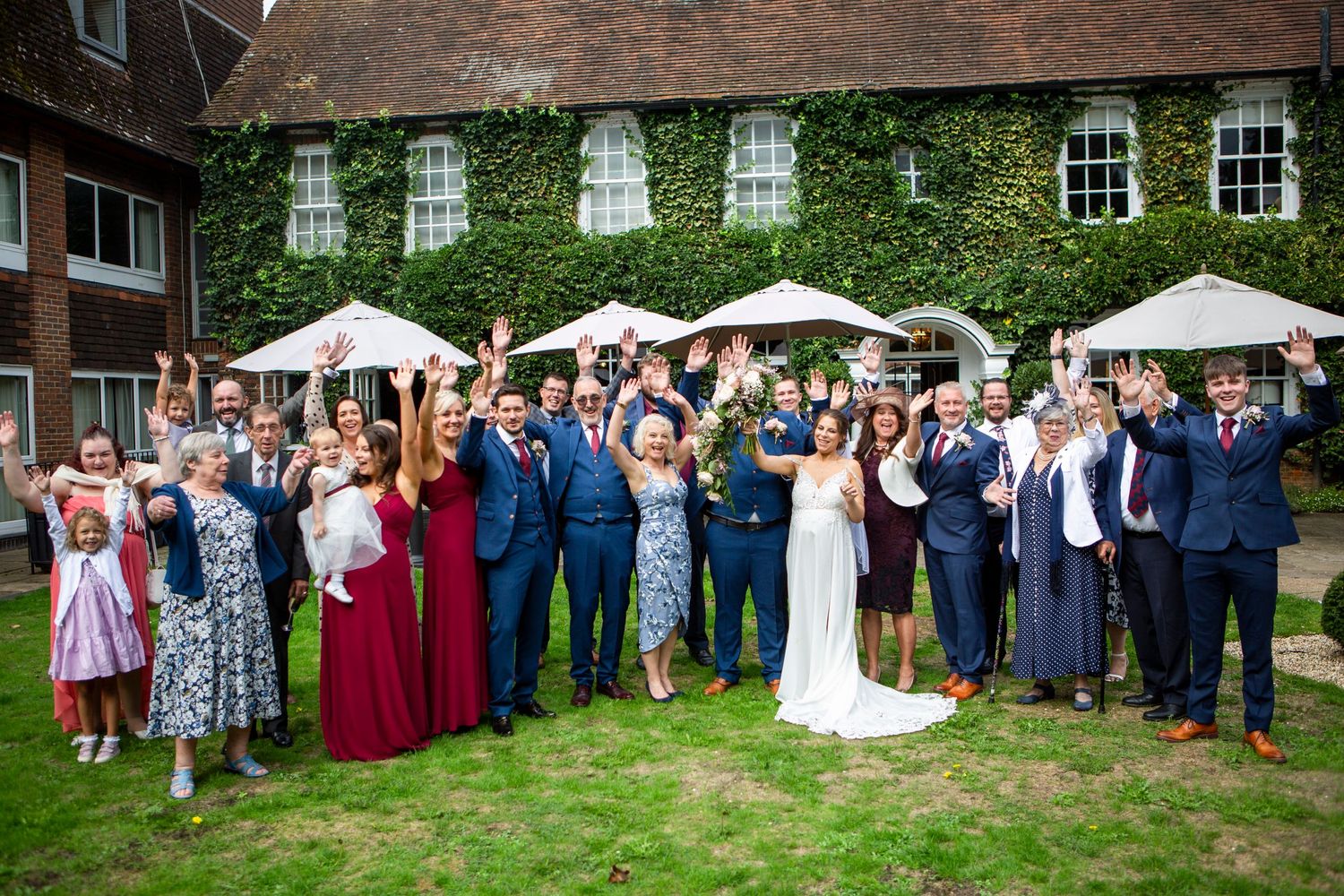 Large wedding party group celebrating outdoors with raised arms in front of ivy-covered venue.