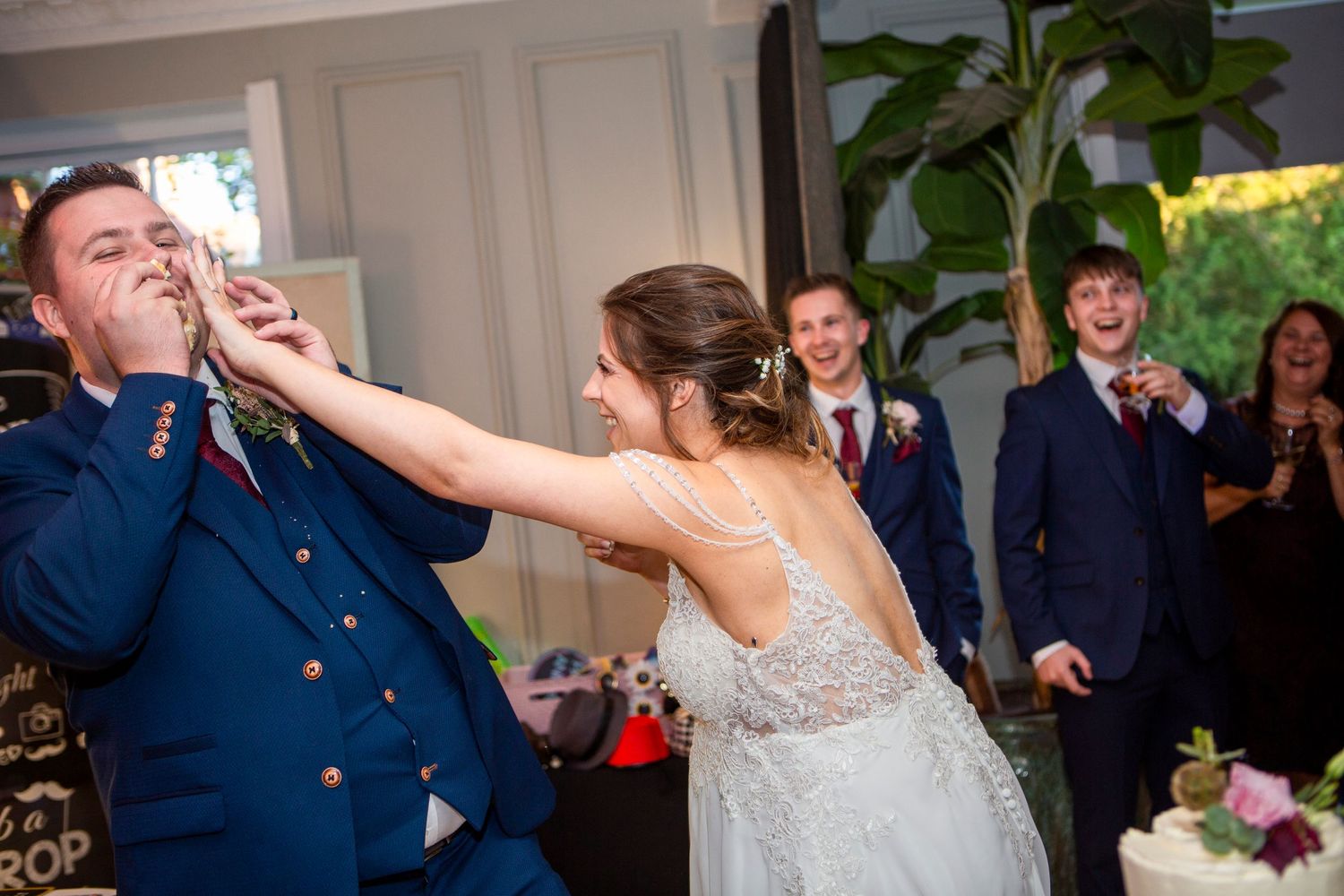 Bride playfully smashing wedding cake into grooms face while guests laugh in background.