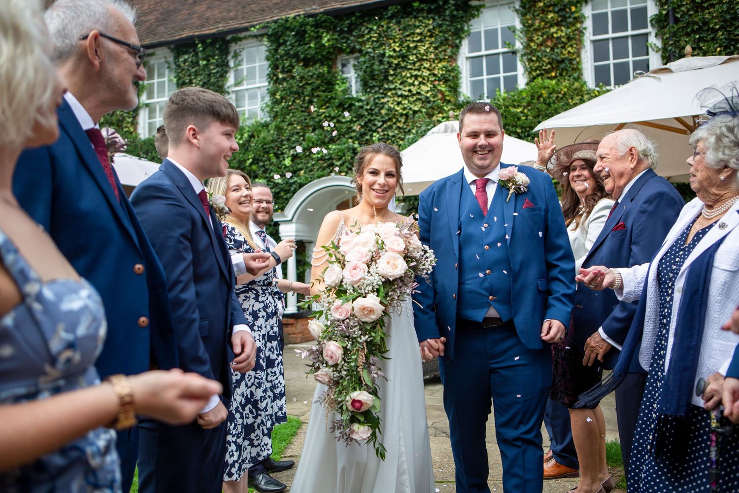 Bride and groom walking through confetti with smiling wedding guests at outdoor reception.
