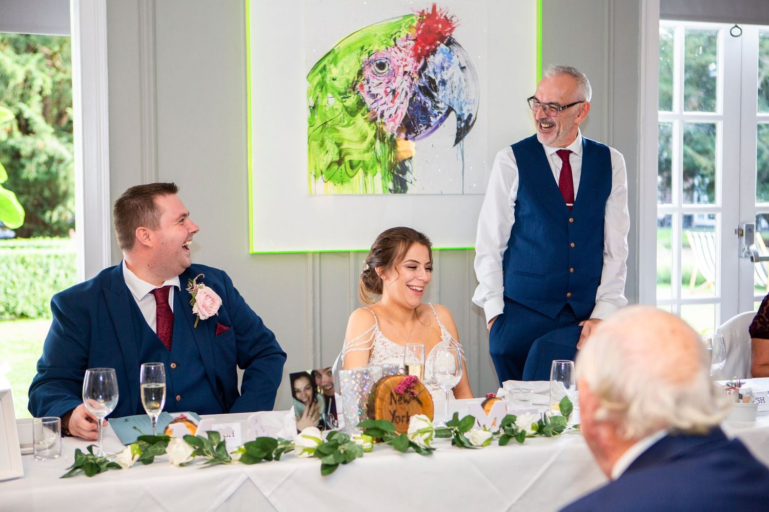 Groom and bride smiling during wedding reception speech at elegantly decorated white table.