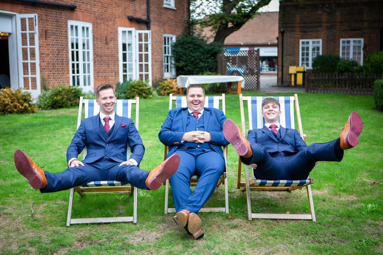 Three groomsmen in navy suits relaxing in striped deck chairs on a lawn.