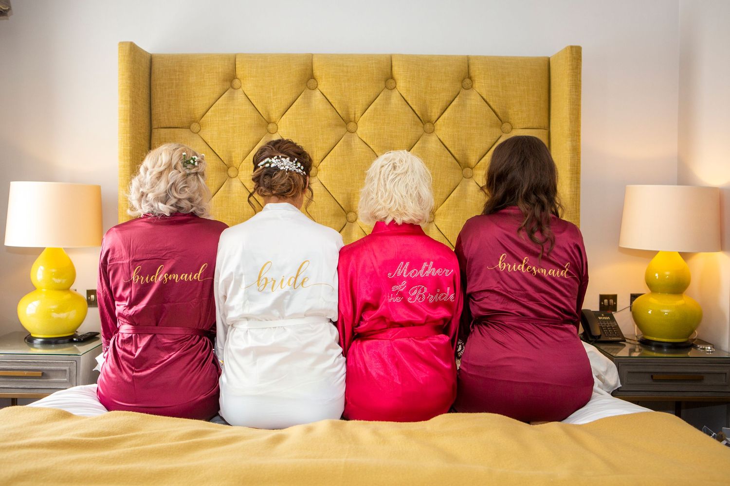 Four women in personalized robes sitting on a bed, showing bridesmaid, bride, mother, and bridesmaid roles.