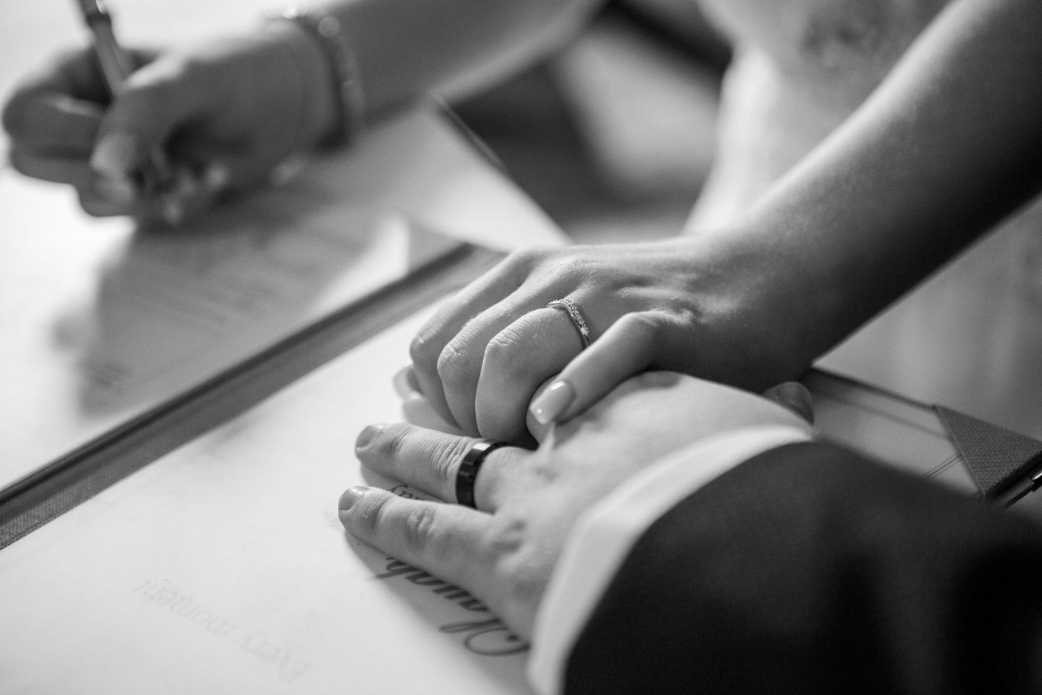 Close-up of bride and groom holding hands while signing wedding register, black and white photo.