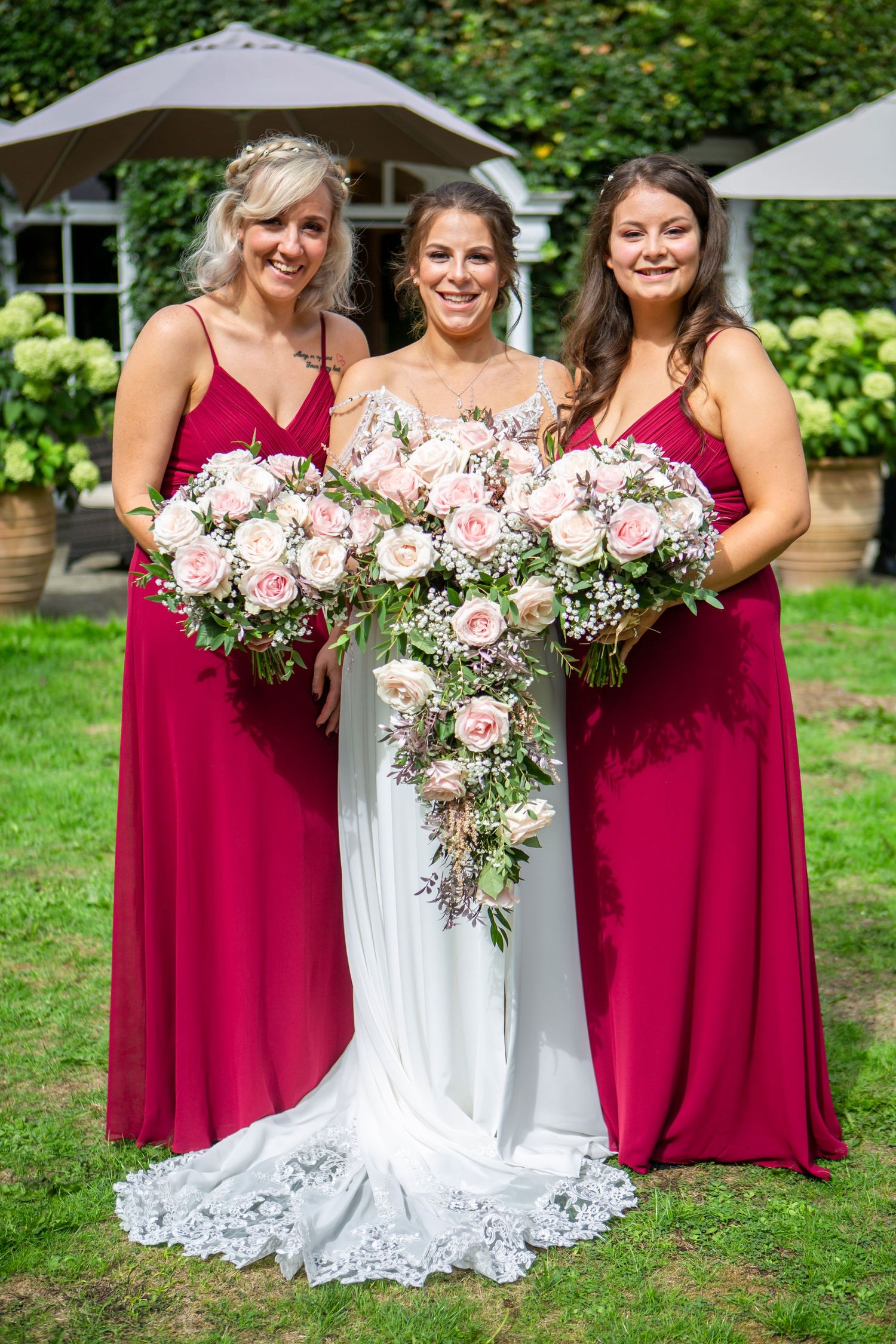Bride flanked by two bridesmaids in red dresses holding beautiful pink rose bouquets outdoors.