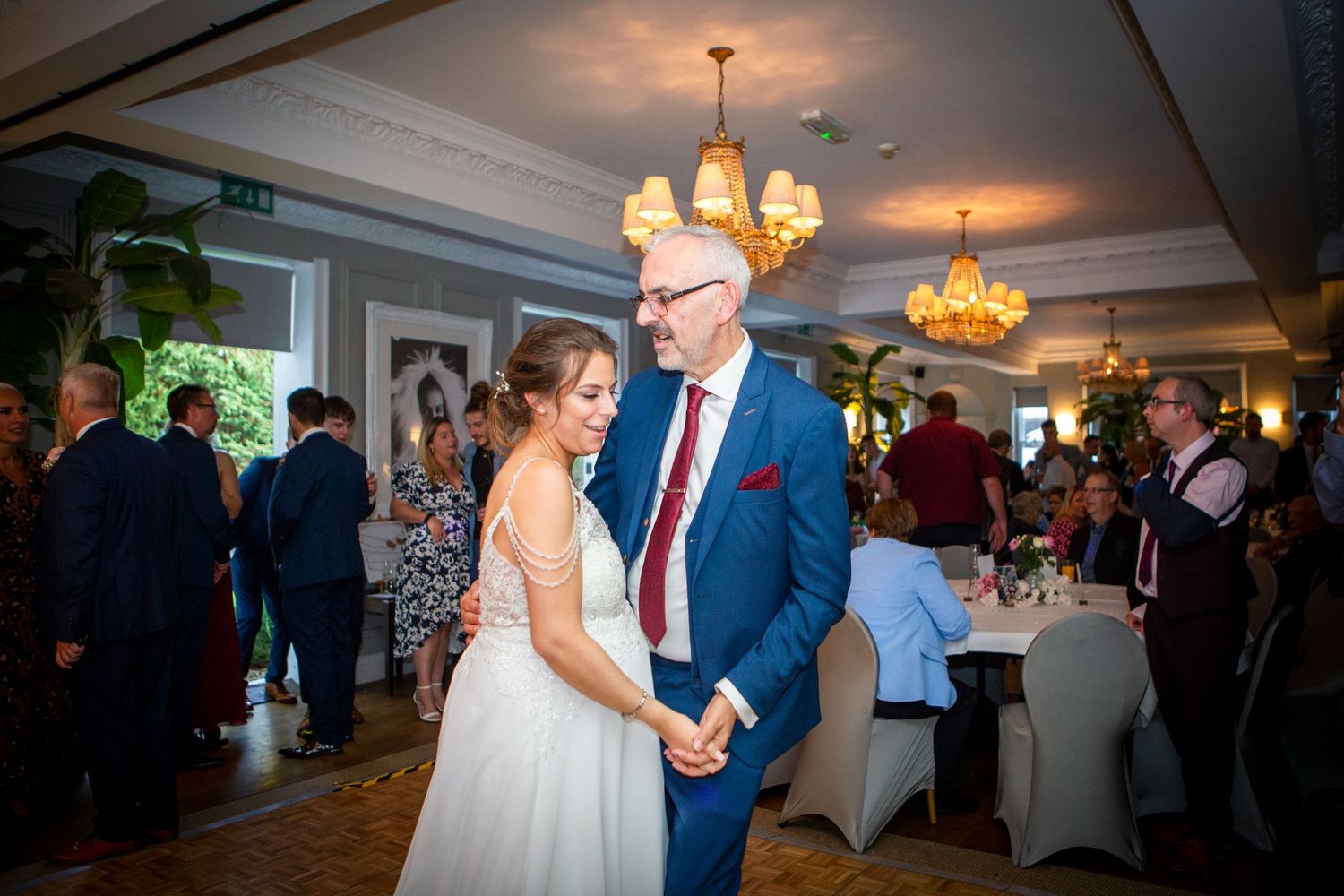 Bride in white gown shares first dance with man in blue suit at elegant wedding reception.