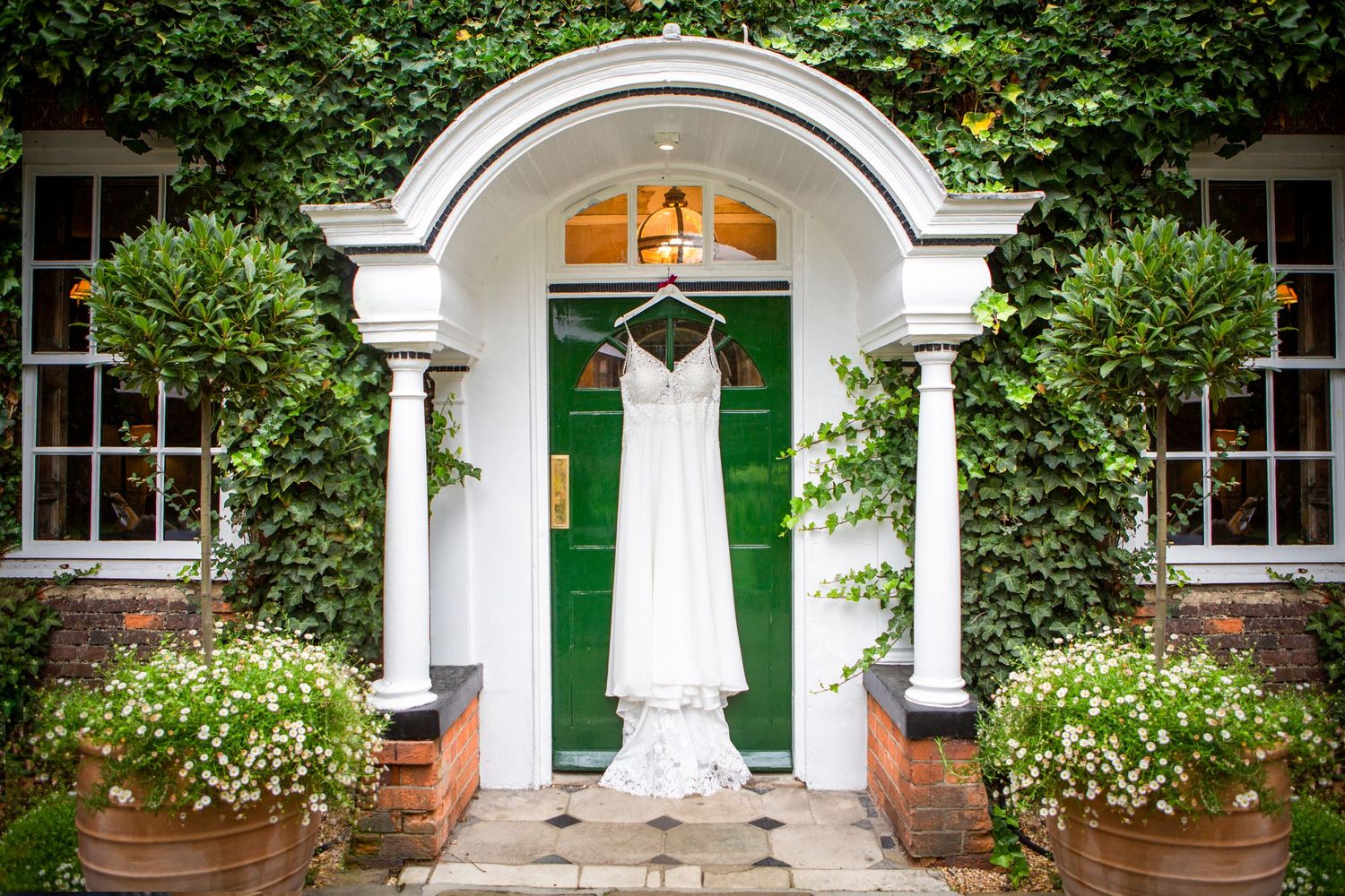Elegant wedding dress hanging on a green door of an ivy-covered building with floral arrangements.
