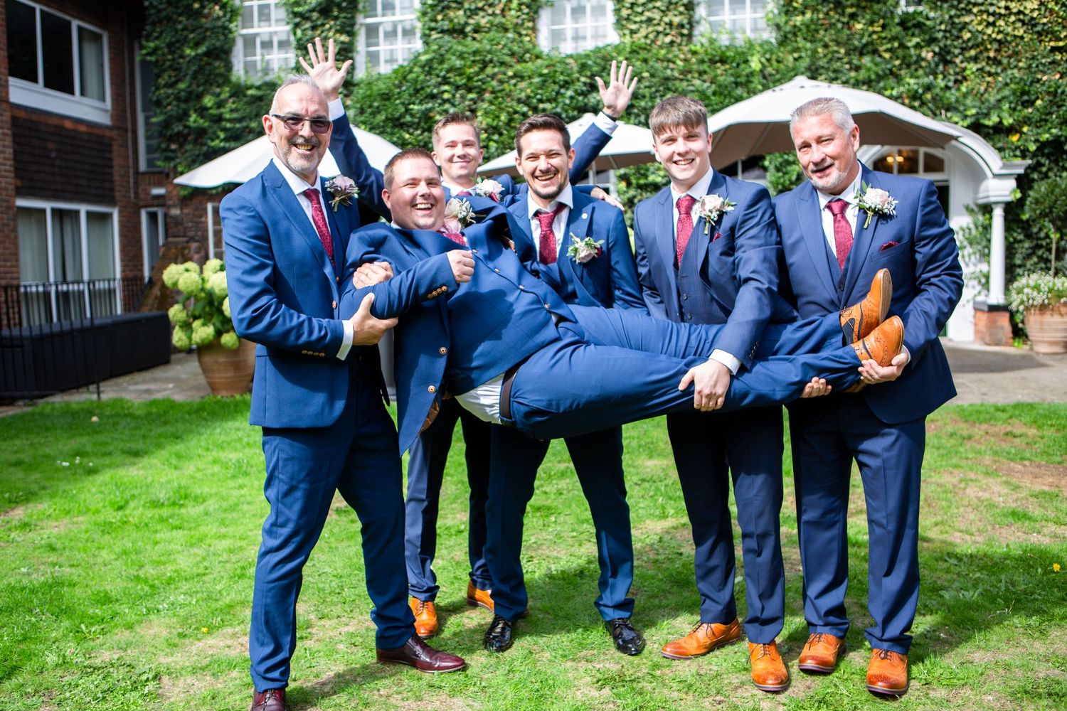 Groomsmen in navy suits lifting groom while celebrating outdoors before wedding ceremony.