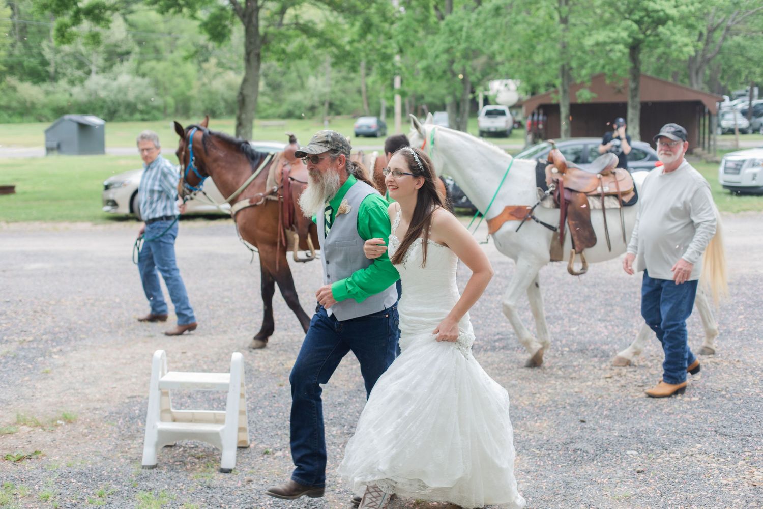 Wedding party members with horses on a gravel path near parked cars.