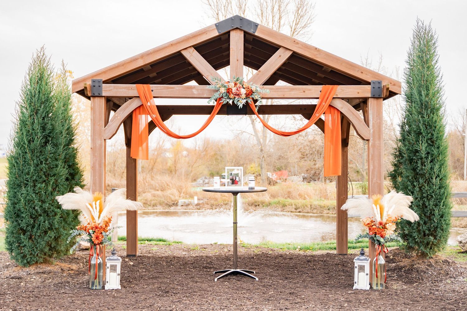 Rustic wooden gazebo decorated with orange ribbons and floral arrangements for an outdoor wedding.