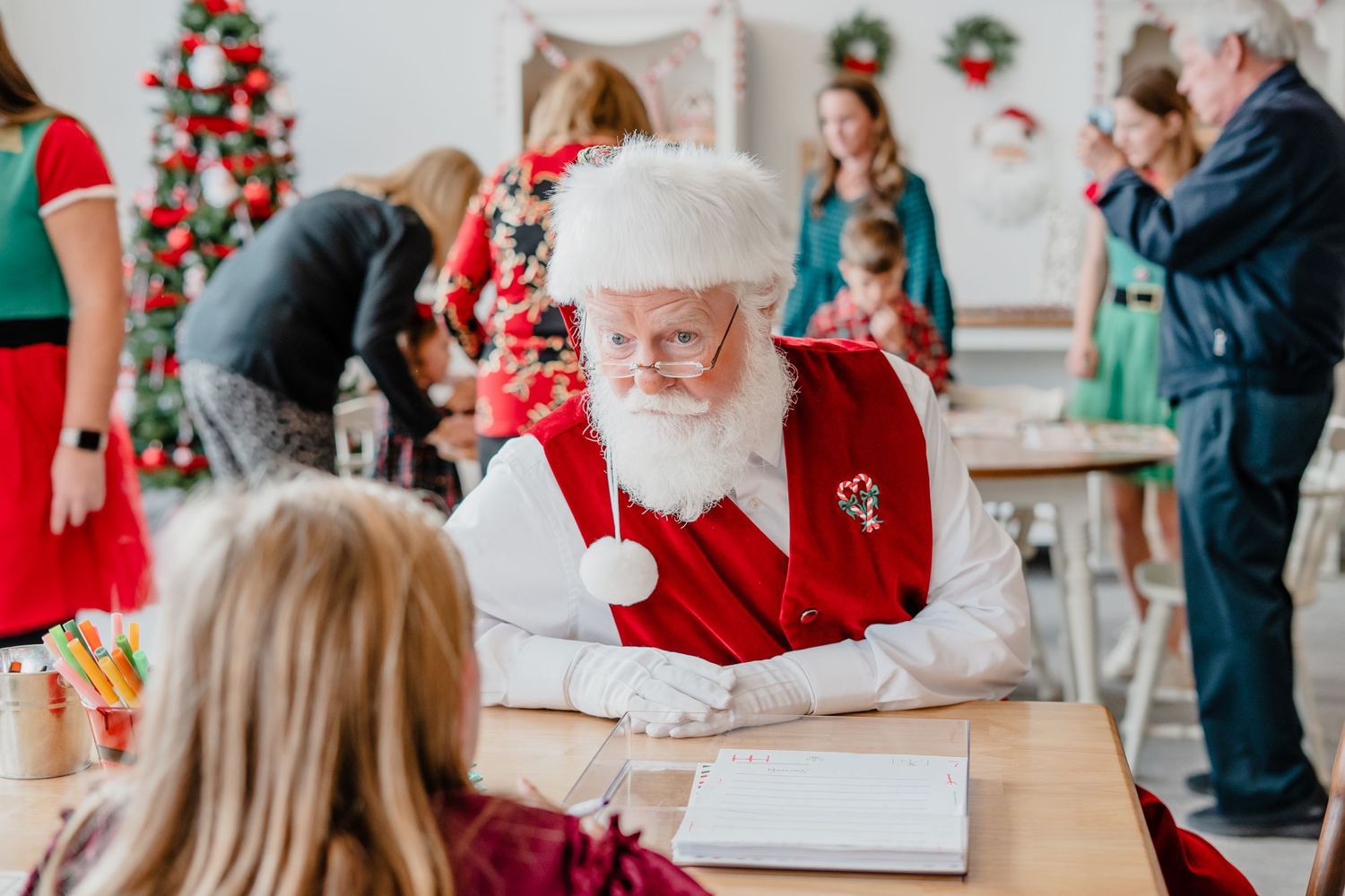 Santa Claus sits at a desk with a book while children and adults gather around during Christmas festivities.
