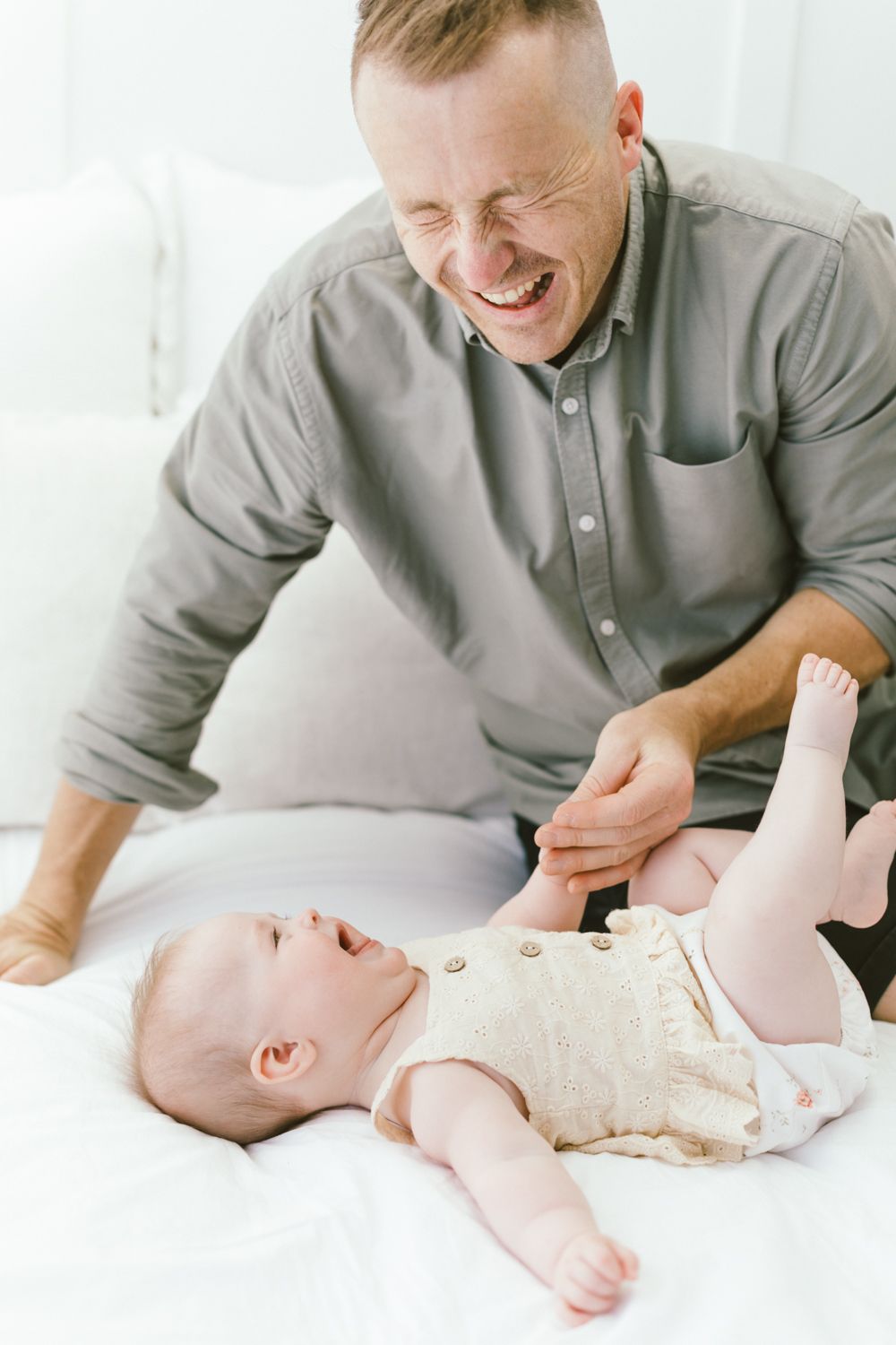 Parent playfully interacting with baby lying on white surface, both sharing joyful expressions.