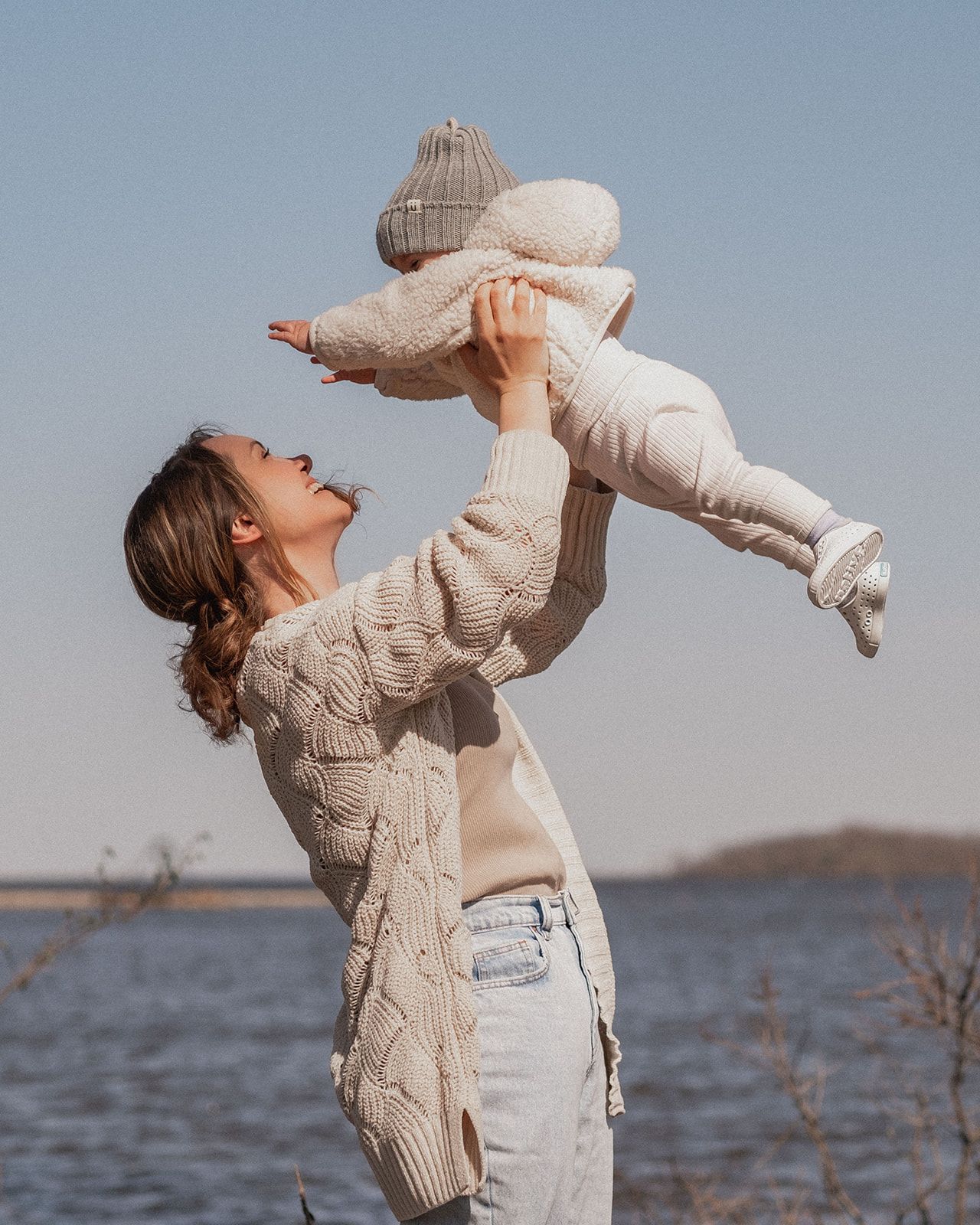 Person in cable knit sweater lifts baby joyfully by waterfront.