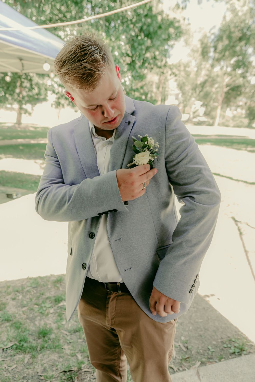 casually dressed blonde groom in grey jacket and chins checking buttonhole before outdoor wedding ceremony