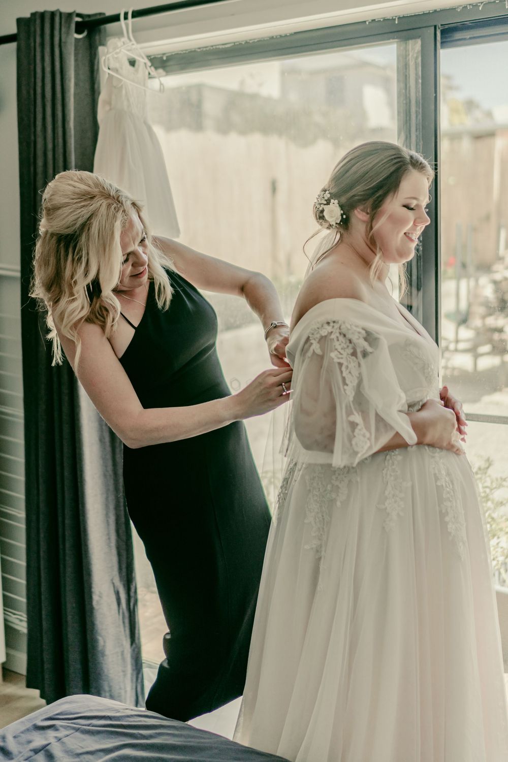 mother of bride in black gown helping pregnant bride in blush dress get ready