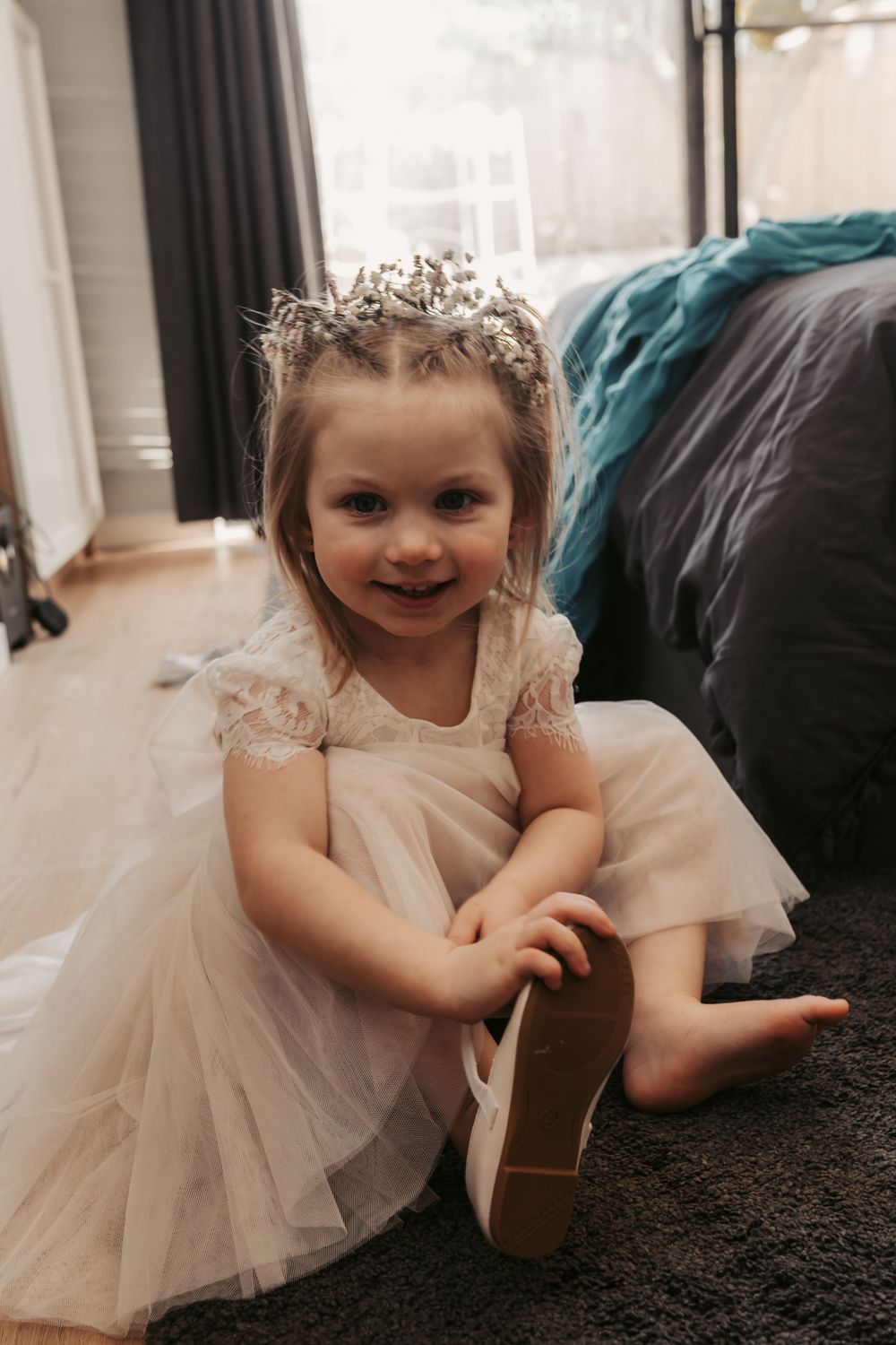 young flower girl in blush dress putting on shoes and smiling at camera