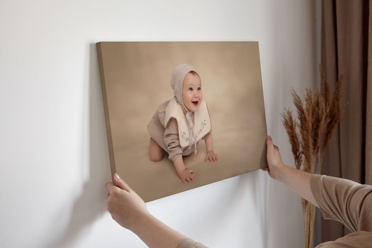 Hands holding up a beige toned portrait canvas against a neutral wall with pampas grass decor.