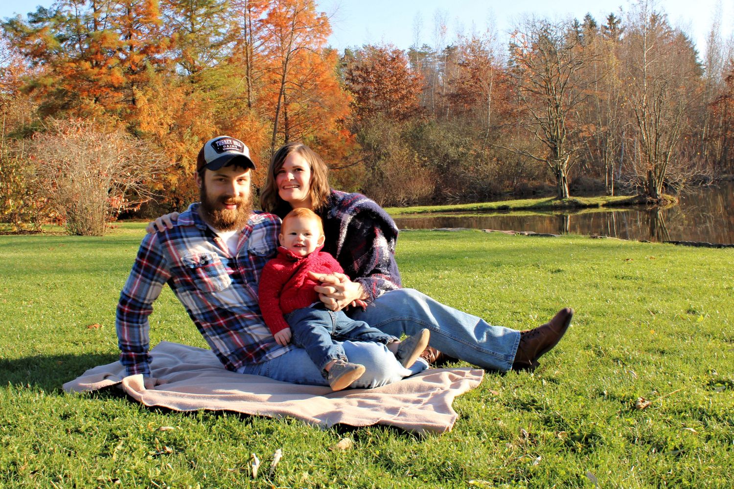 A family of 3 sitting on the grass on infront of a pond on a fall evening.