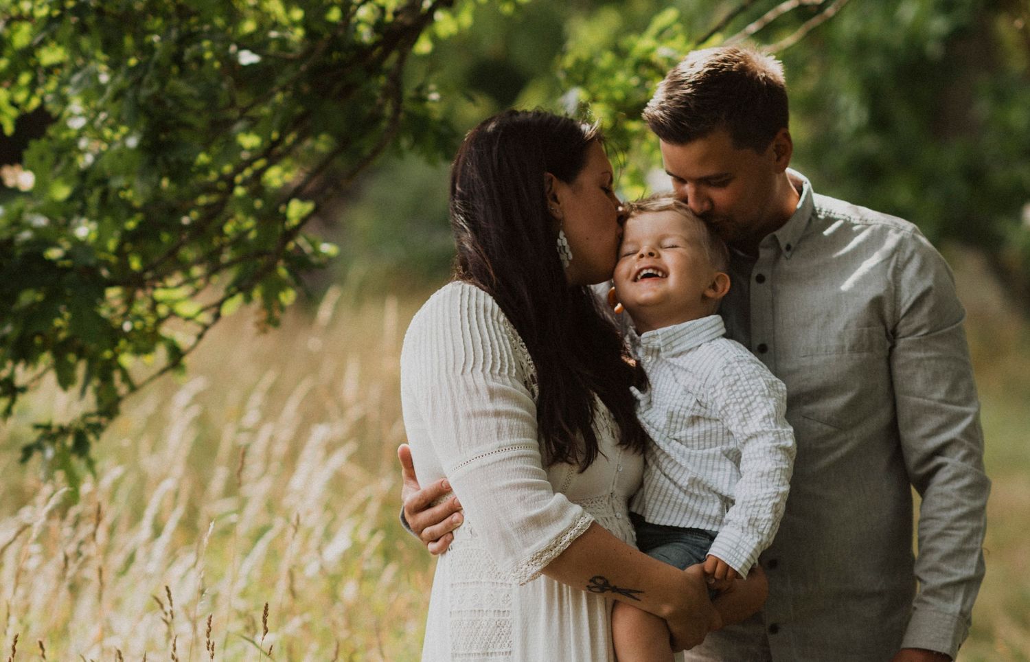A happy young family with a toddler sharing a tender moment outdoors in a sunlit meadow with lush green trees.