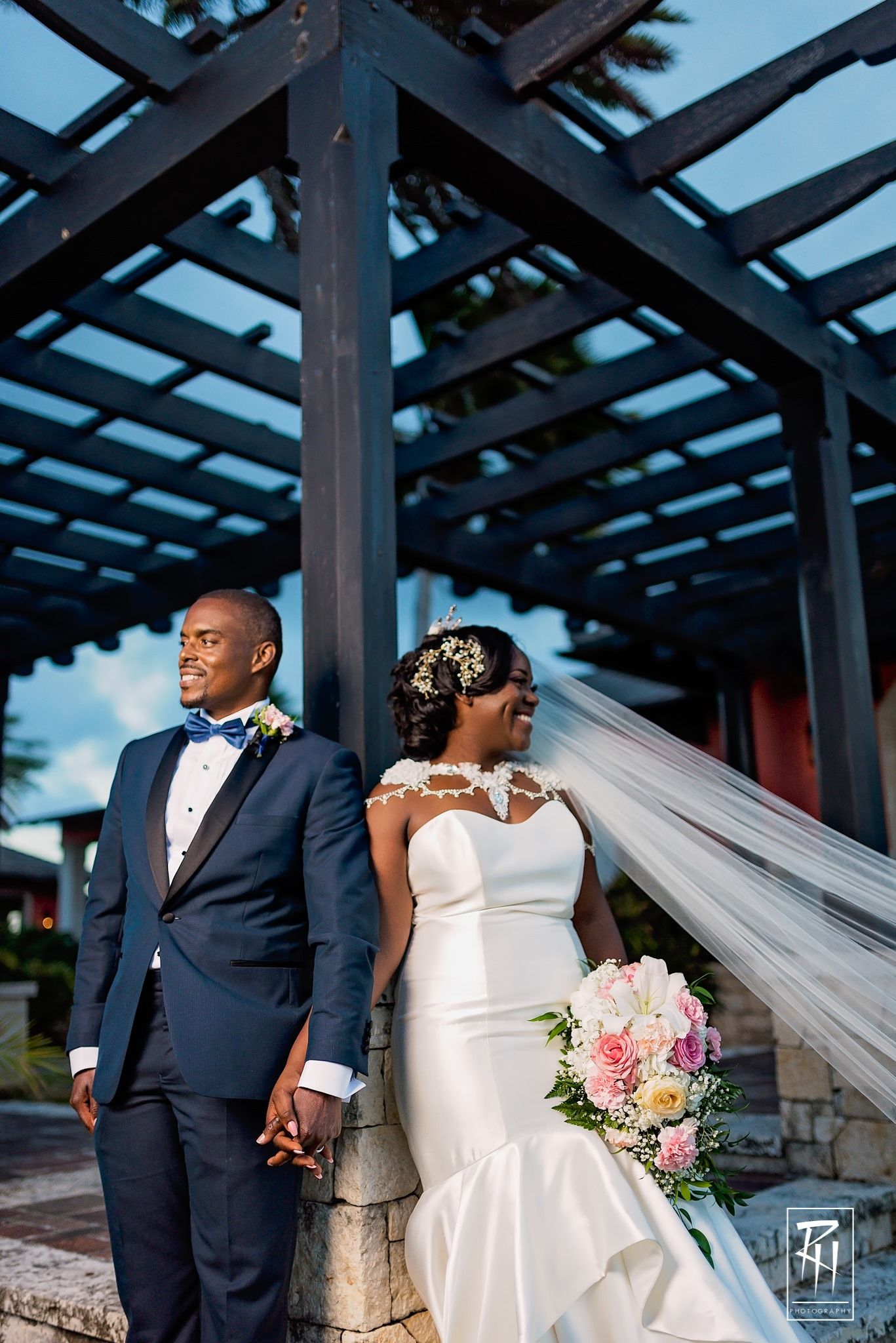 Elegant wedding couple poses under pergola with flowing veil and pink bouquet.