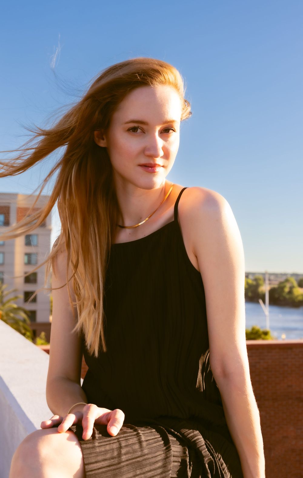 A black tank top contrasts against warm sunlight with a river and buildings in the background.