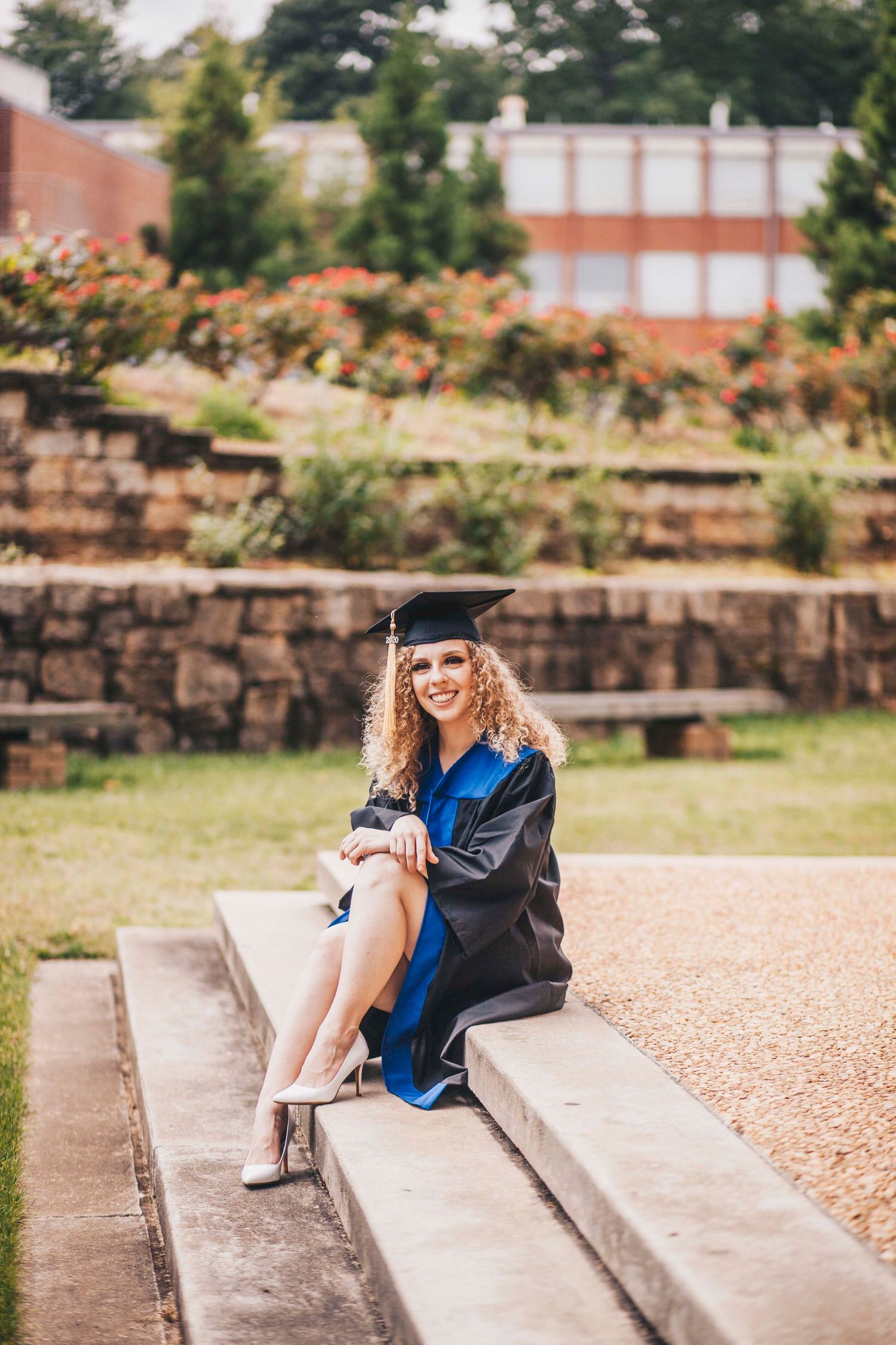 Smiling female graduate in cap and gown sitting on stone steps in campus garden.