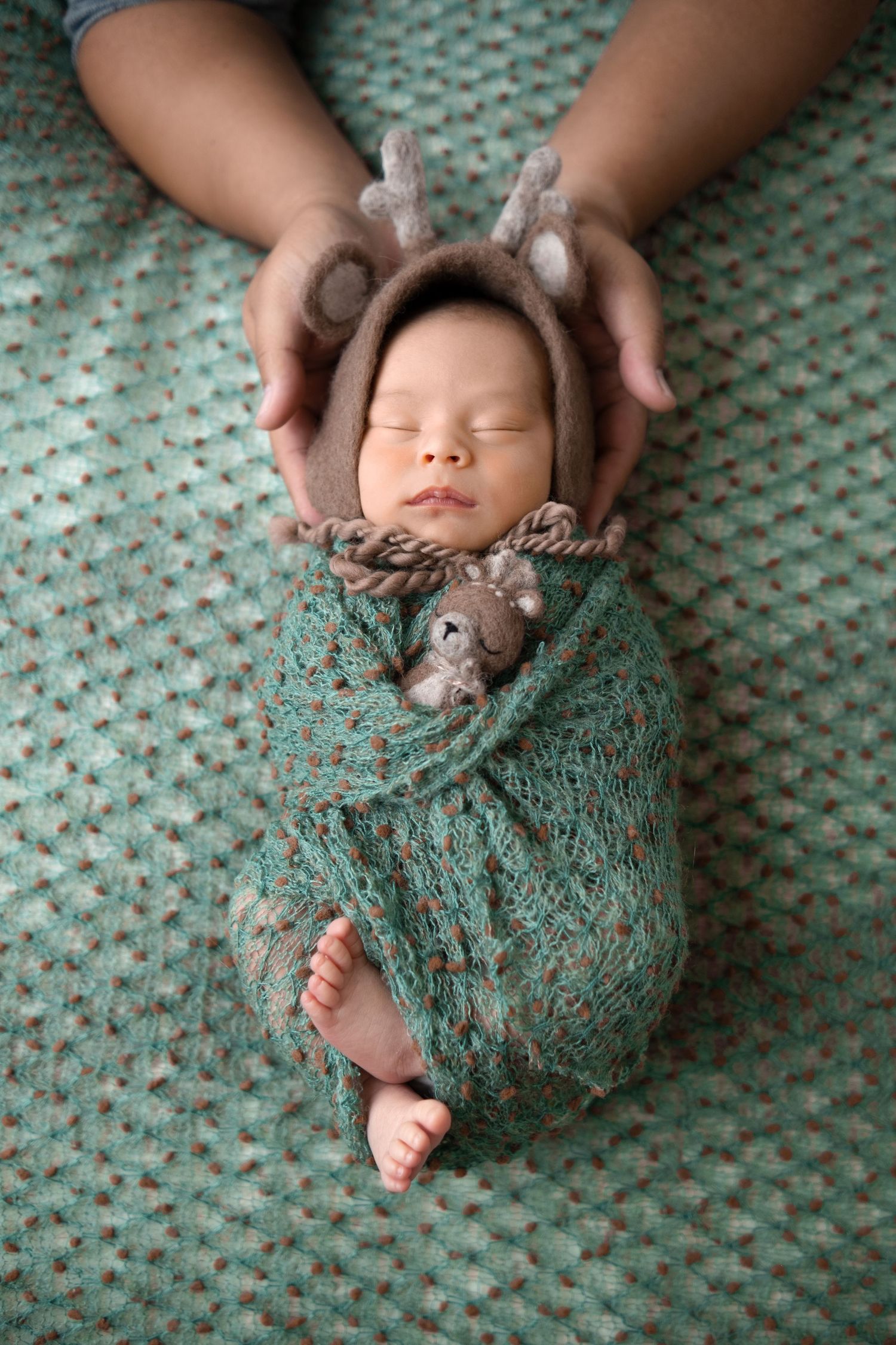 Sleeping baby wrapped in teal knitted blanket wearing reindeer hat and holding plush toy on textured background.