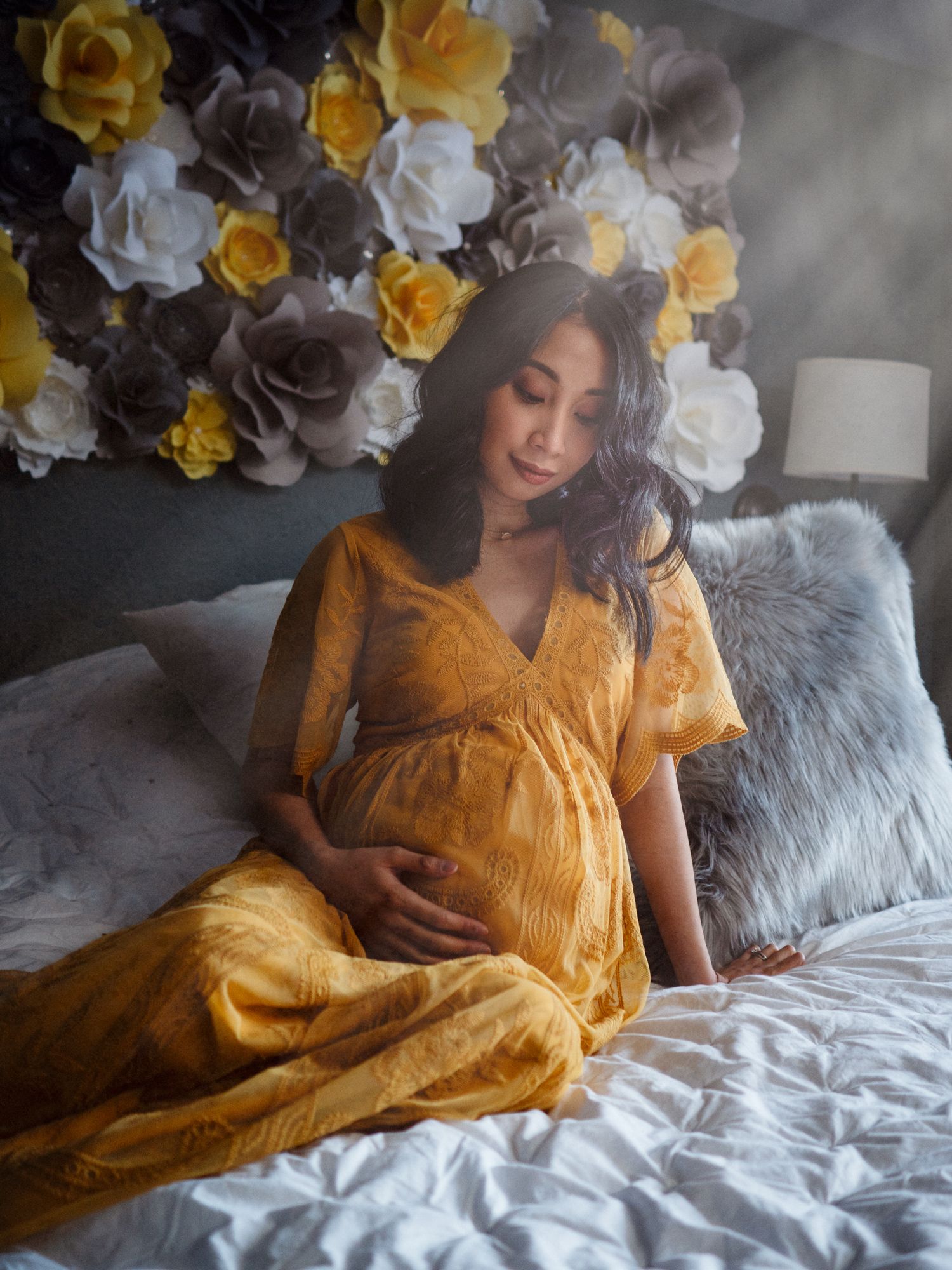 A pregnant woman in a mustard yellow dress relaxes on a bed with gray pillows under white and yellow paper flowers.