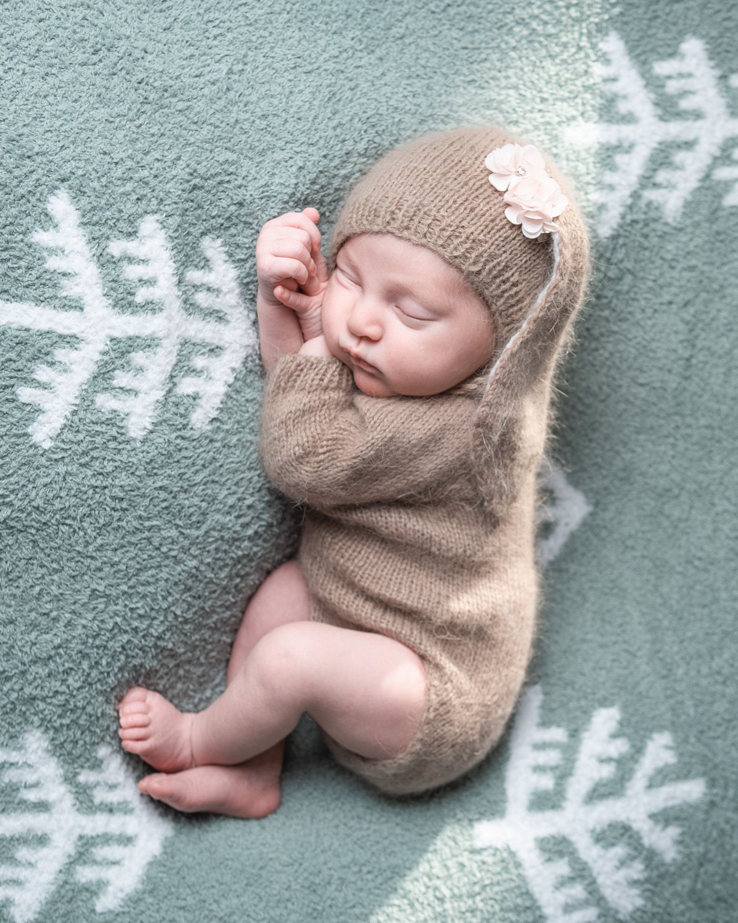 Sleeping baby wearing cozy brown knit sweater and beanie on teal blanket with snowflake pattern.