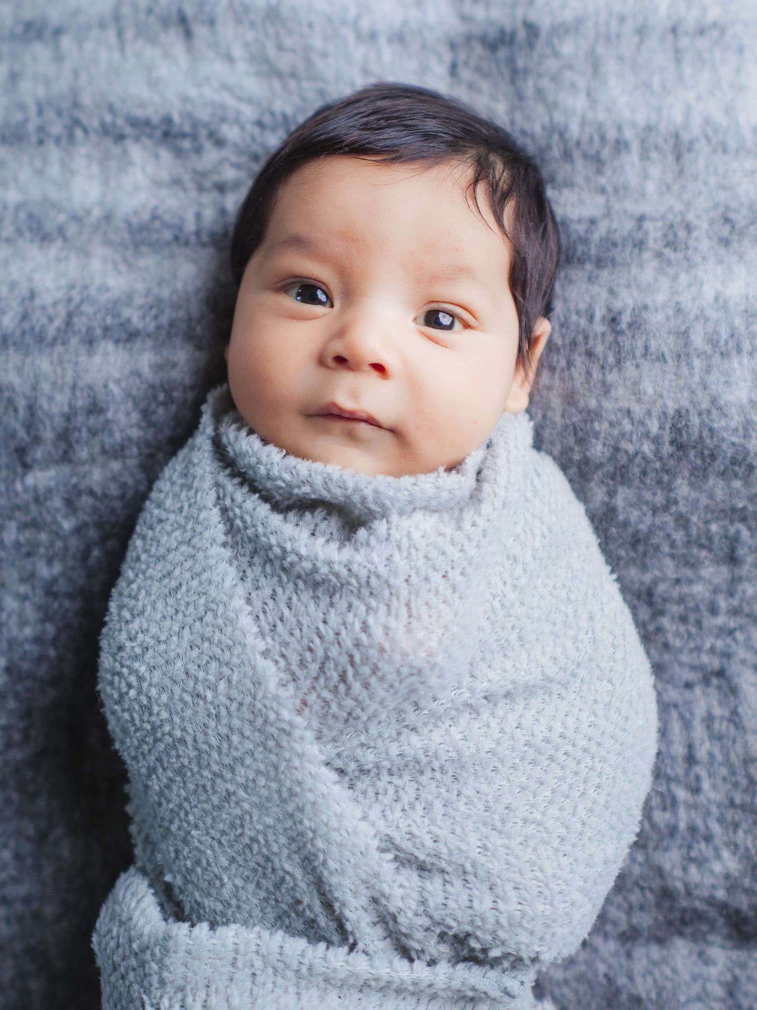 A baby wrapped in a cozy gray knit blanket lies on a textured gray background looking up at the camera.