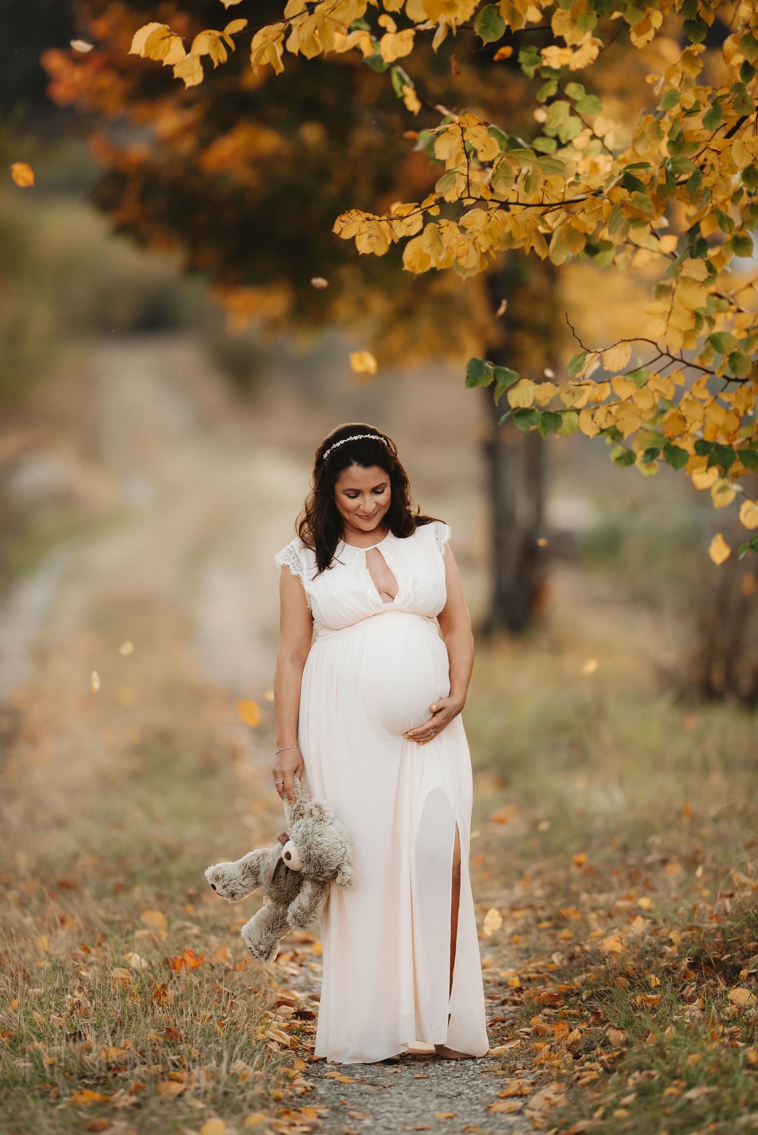 A maternity portrait under golden autumn leaves with a white dress and small plush toy.
